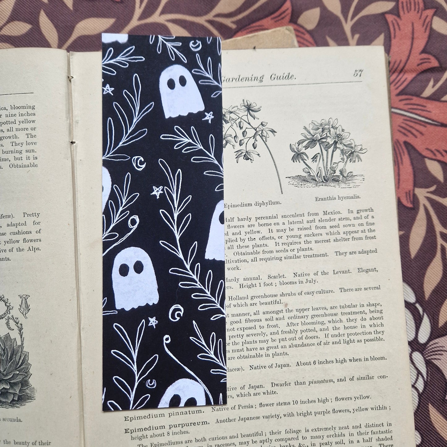 An open vintage gardening book sits on a brown floral patterned background. The book has a black bookmark featuring a white line pattern of ghosts among a leafy vine with stars and crescent moons.
