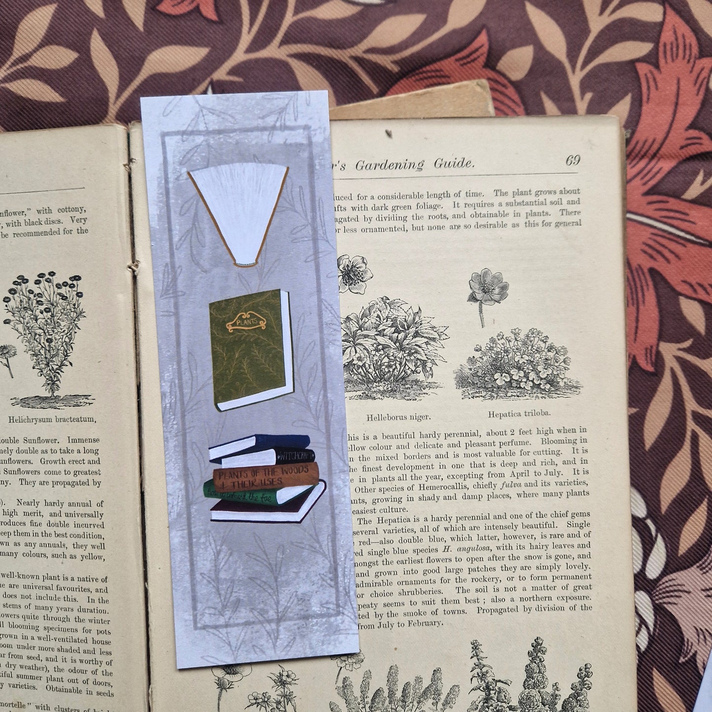 The front of the bookmark is visible in an open vintage gardening book on a brown retro floral patterned fabric. The bookmark is a textured grey with a darker grey border line running around the outside as well as a leafy vine design. Running down the bookmark are three different book illustrations - a purple book, a stack of books and then a green book.