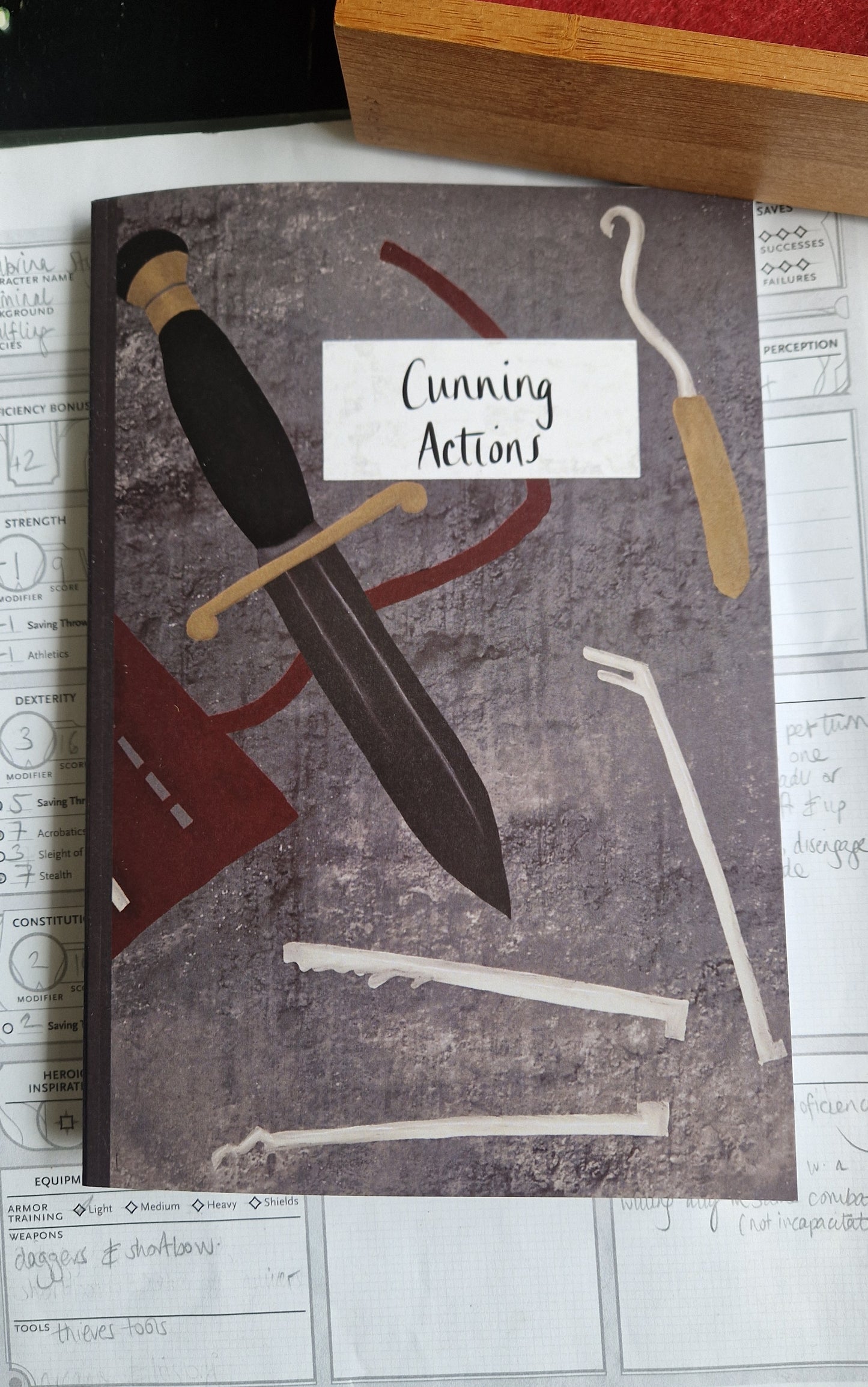 The Reckless Attack Notebook by Duck Egg Designs, made from recycled paper and featuring a dagger, tools, and a red pouch on the cover, sits atop a TTRPG character sheet—perfect for jotting down campaign notes.