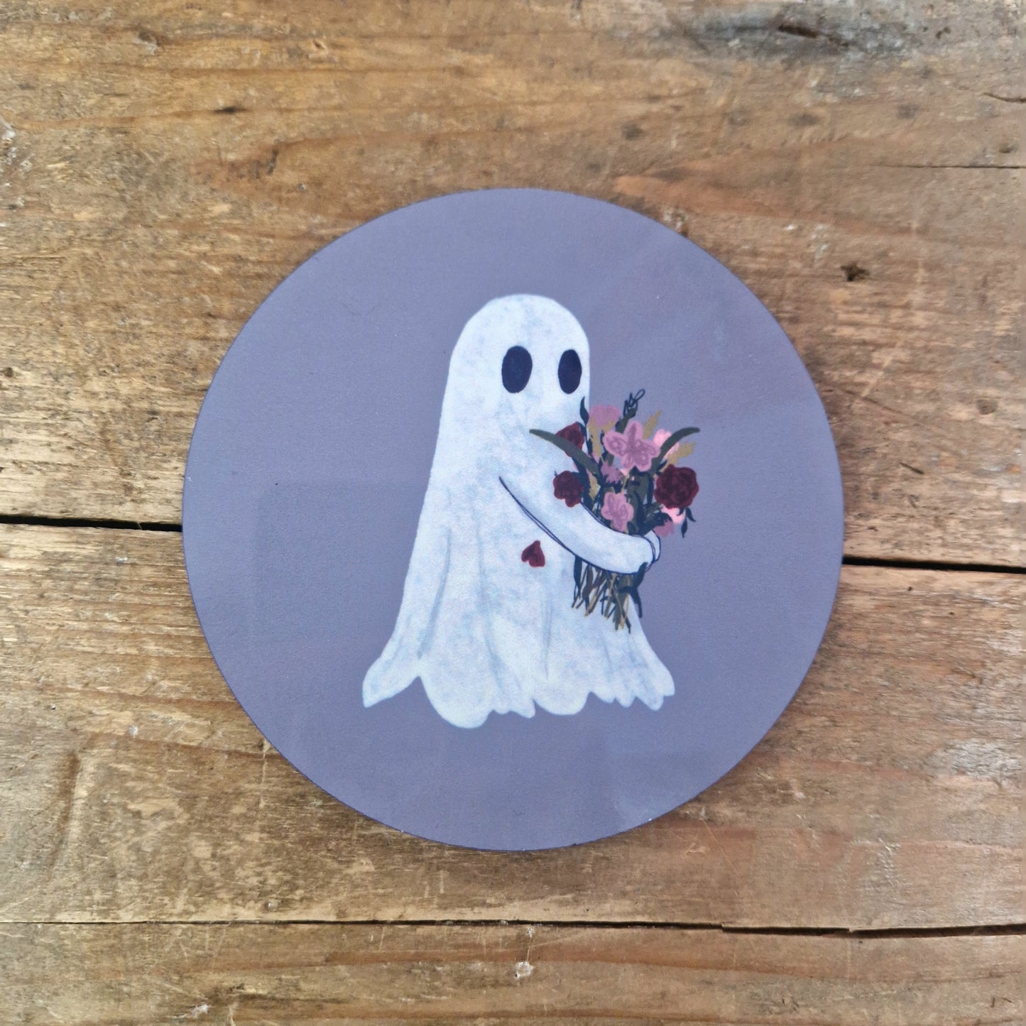 The Flower Ghost Coaster by Duck Egg Designs features a cartoon ghost with a bouquet and red heart on its chest, set against a purple circle. UK printed, this charming coaster adds whimsy to any wooden surface.