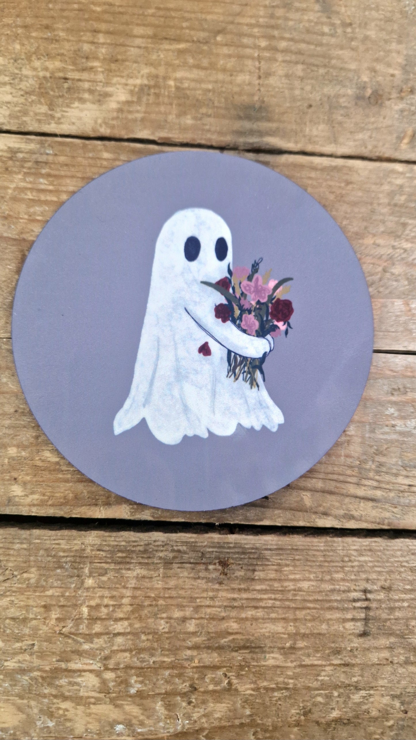 The Flower Ghost Coaster by Duck Egg Designs features a cute cartoon ghost holding a colorful bouquet on a grayish oval background, shown on a rustic wooden surface.