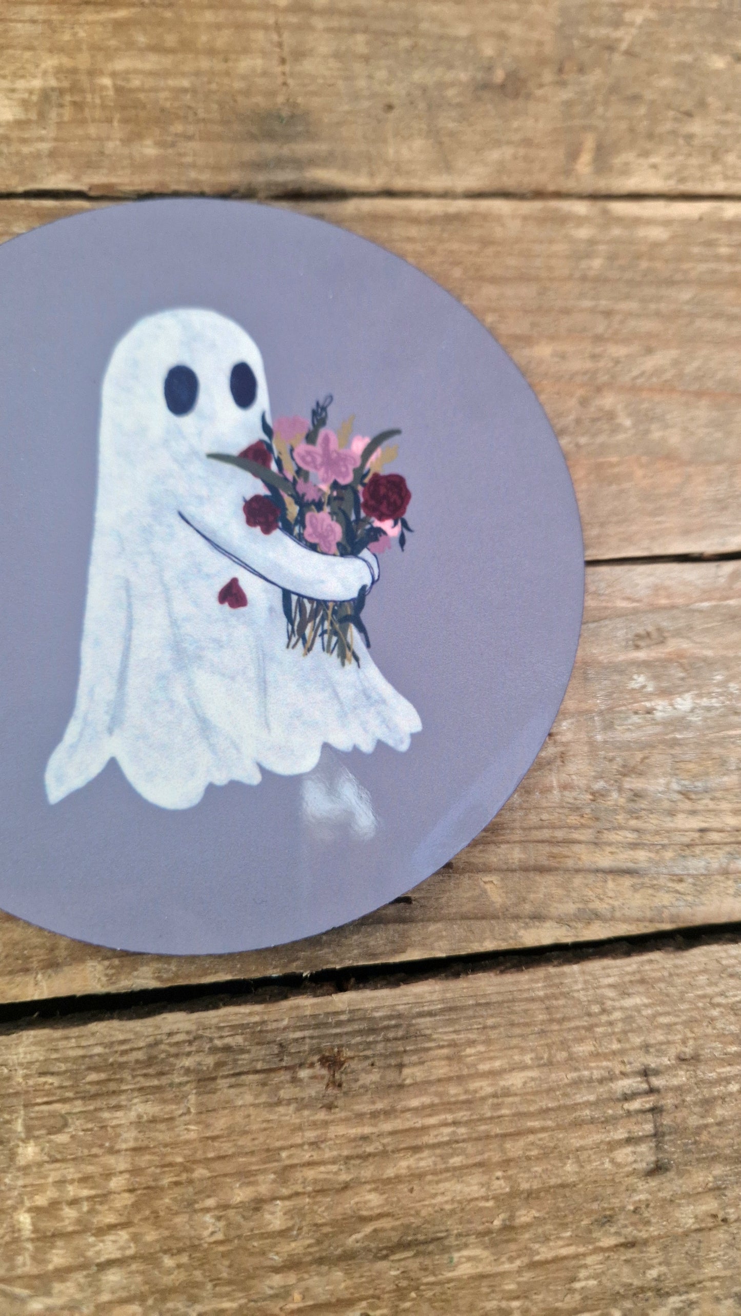 A sticker inspired by Duck Egg Designs' Flower Ghost Coaster features a cute cartoon ghost with a colorful bouquet on a purple background, set on a wooden surface.