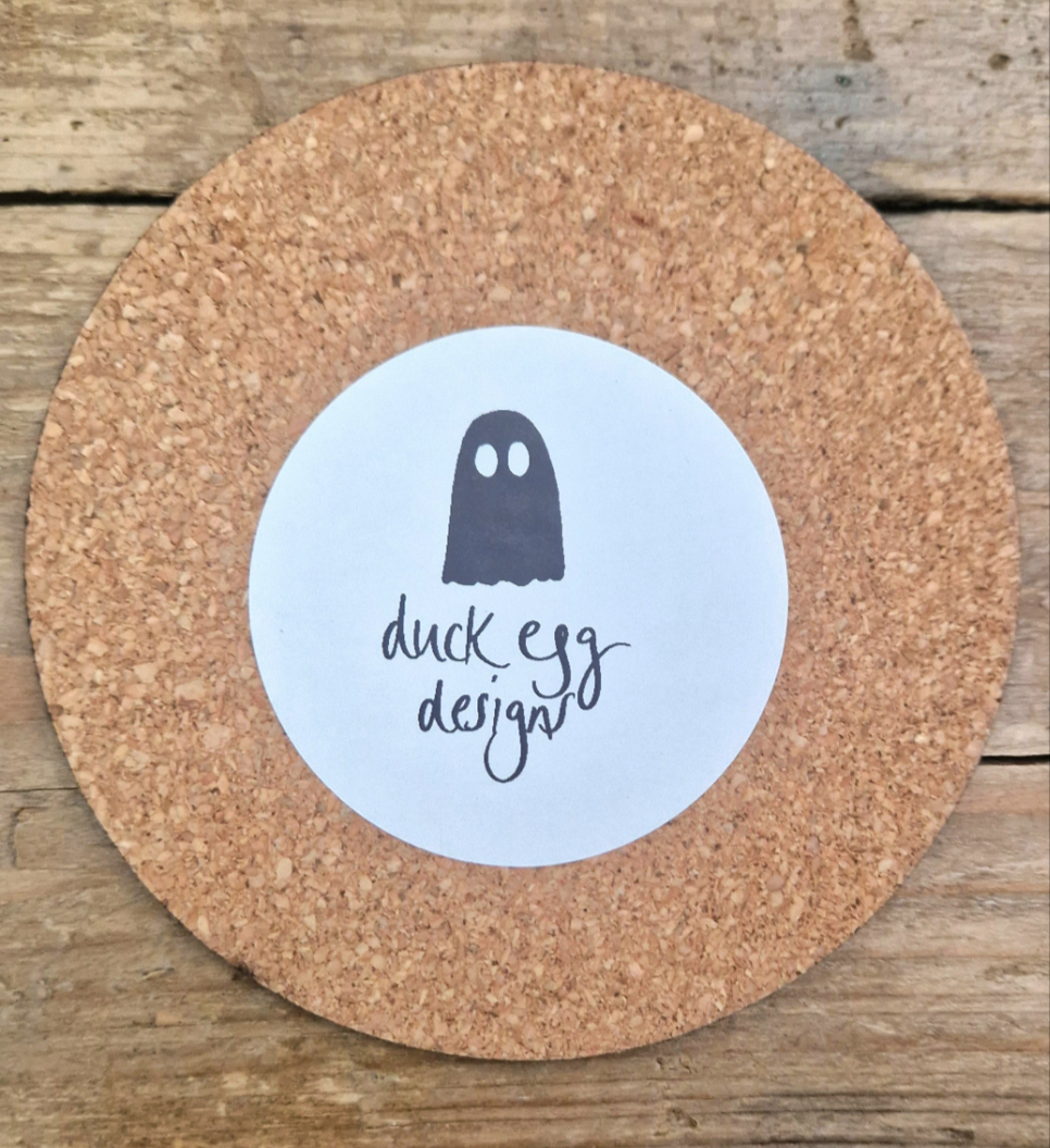 The Obsessed With Cats Coaster by Duck Egg Designs features a round cork base and white label with a simple black cat illustration, printed in the UK, and is shown resting on a wooden surface.