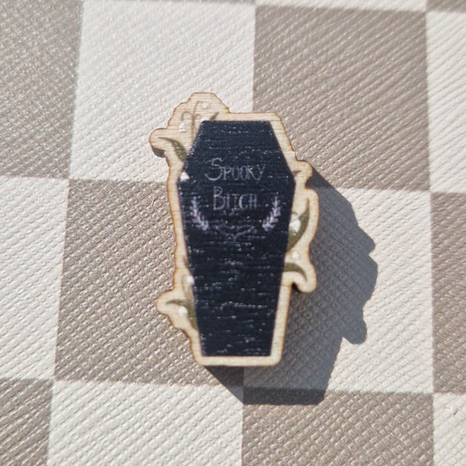A small wooden magnet with a 'Spooky Bitch' inscription, designed to resemble a coffin, laid on a checkered surface.