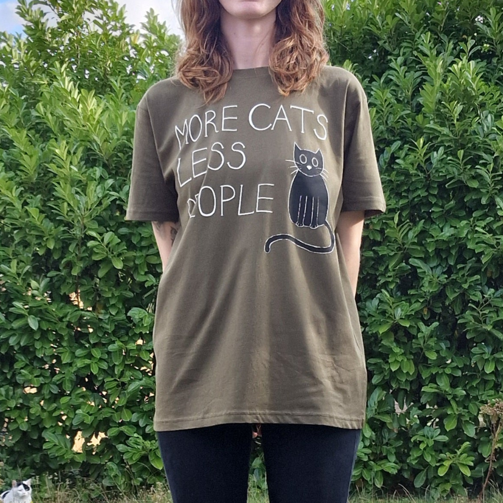 A person stands in Norwich by green bushes, wearing Duck Egg Designs’ More Cats Less People Tshirt—an olive green tee with "MORE CATS LESS PEOPLE" and a black cat drawing.