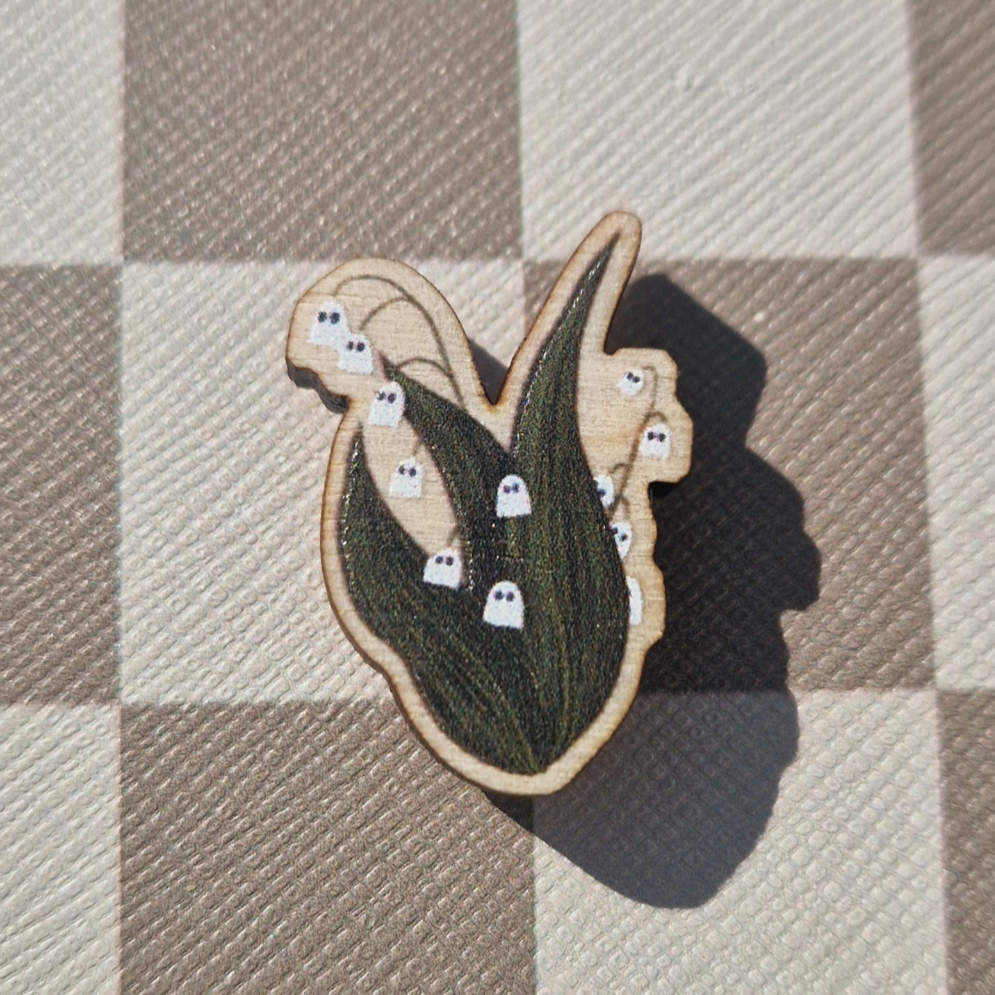 A wooden magnet in the shape of a lily of the valley with a ghost pattern, placed on a checkered surface.