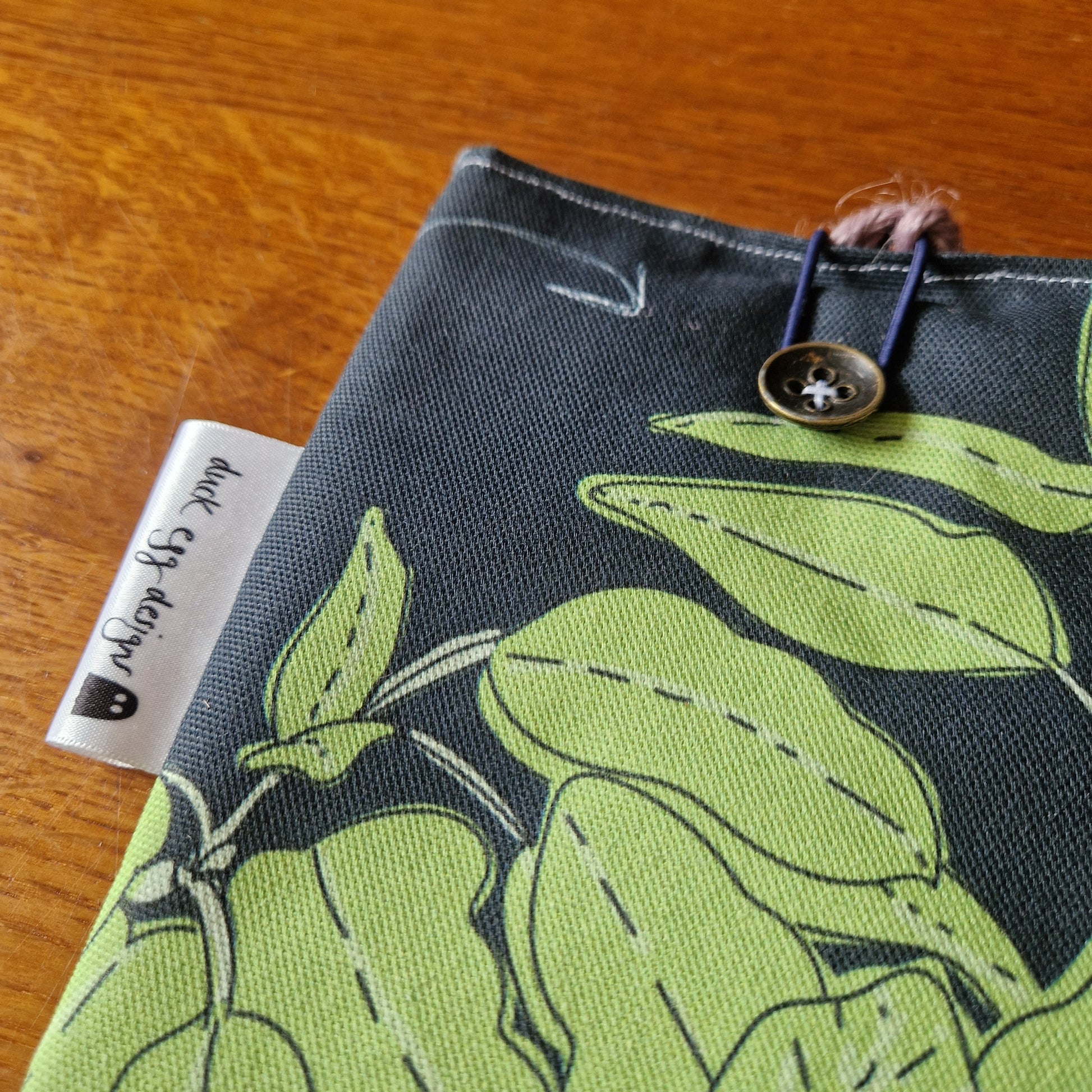 The Common Herbs Kindle Pouch by Duck Egg Designs features a green leaf pattern, a button and elastic loop closure, and a sewn-in brand tag on the side seam. This handmade sleeve is displayed on a wooden surface.