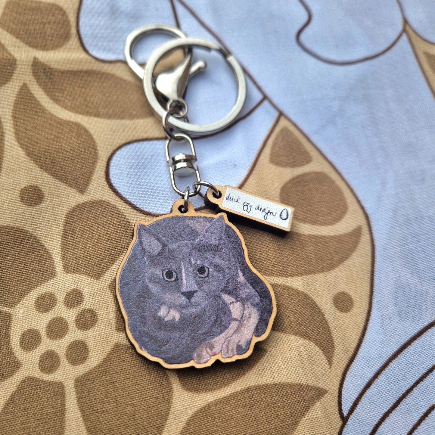 The Luna Grey Cat Keyring by Duck Egg Designs is a UK-made keychain featuring an illustrated grey cat lying down and a small metal tag engraved with "luck off design." It is displayed on patterned brown and blue fabric.