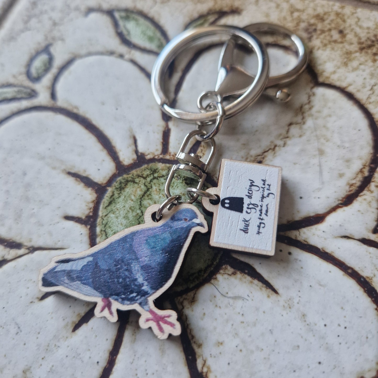 The Paloma Pigeon Keyring by Duck Egg Designs features a painted pigeon charm and a small rectangular tag with handwritten text, shown on a floral ceramic surface.