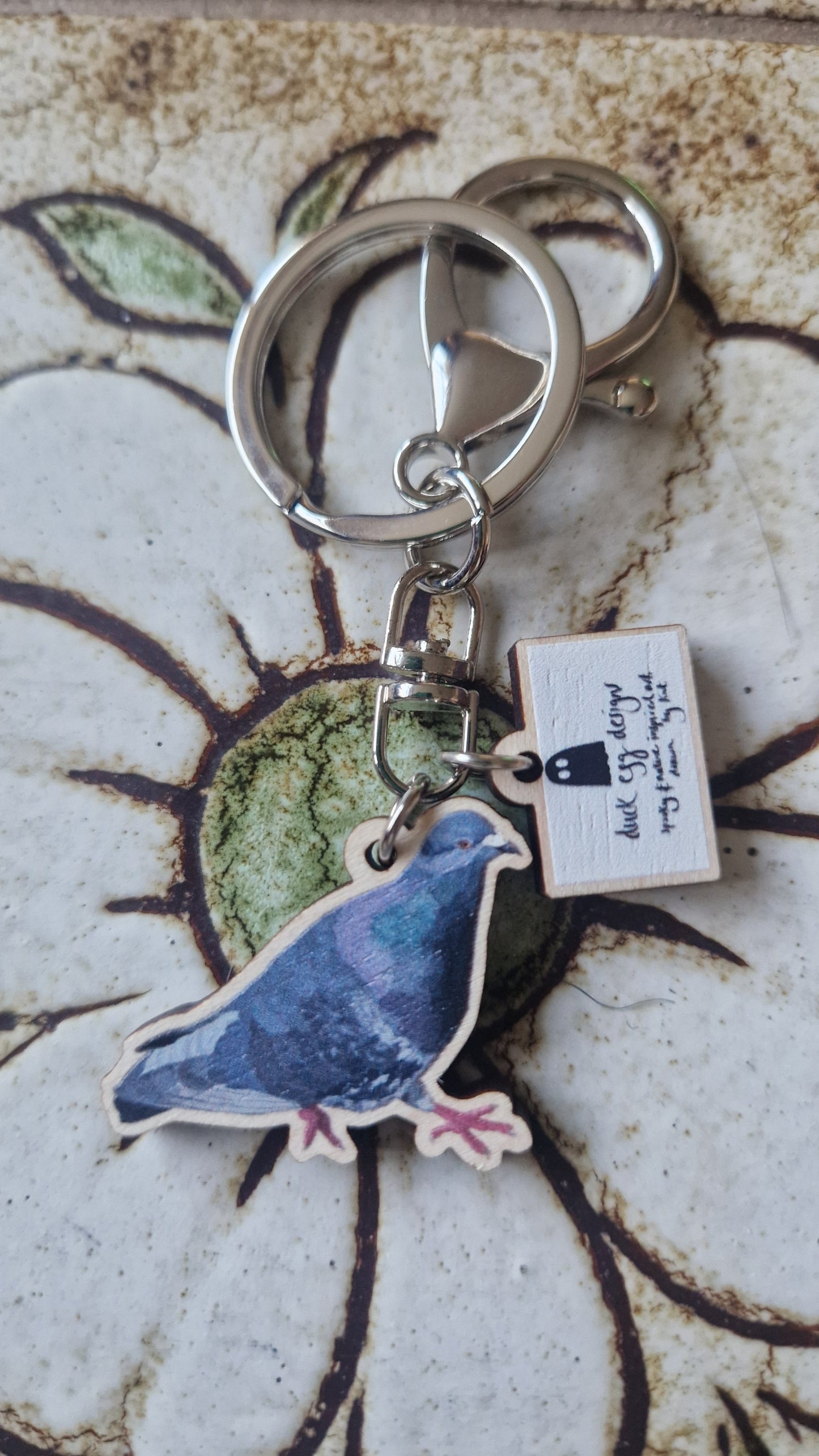 The Paloma Pigeon Keyring by Duck Egg Designs features a cutout pigeon illustration and a small tag reading “Mr. Gregory the homing pigeon,” displayed on a textured, flower-patterned tile.