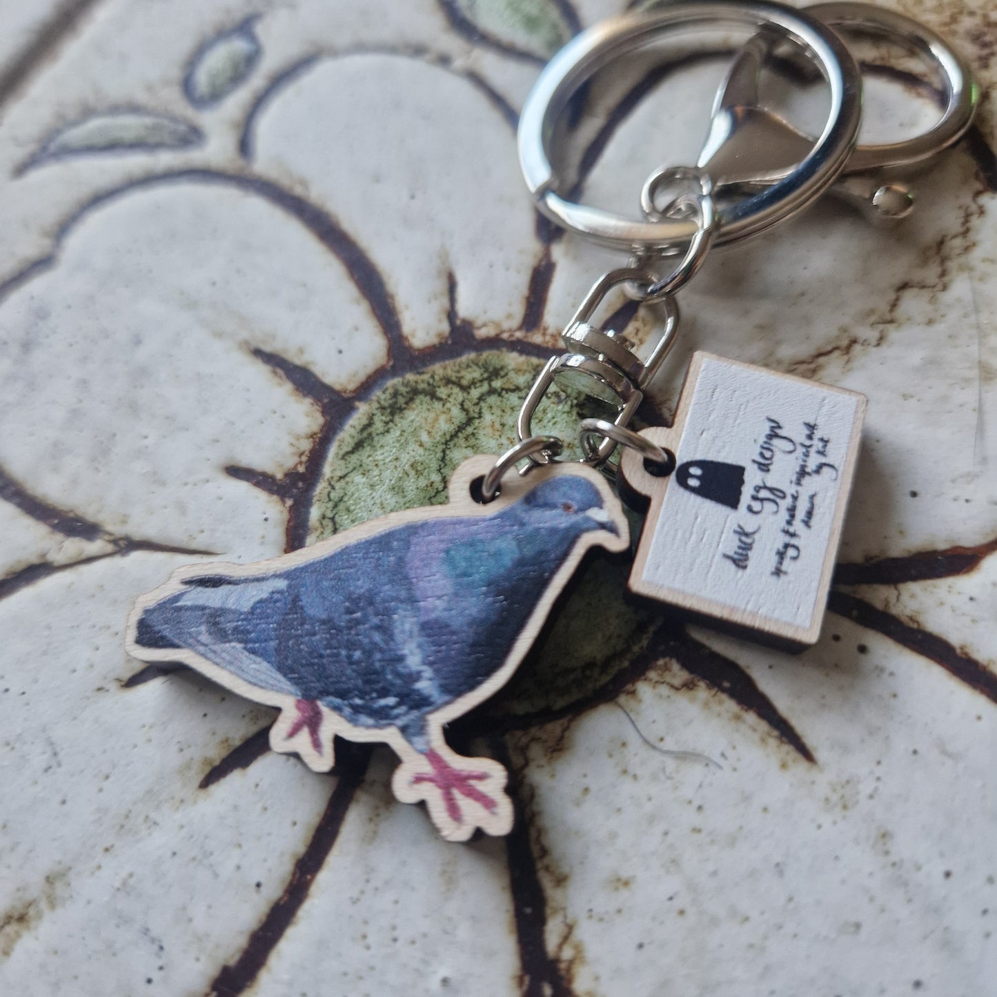 The Paloma Pigeon Keyring by Duck Egg Designs features a pigeon charm and a small rectangular tag with handwritten text, displayed on a floral ceramic surface.