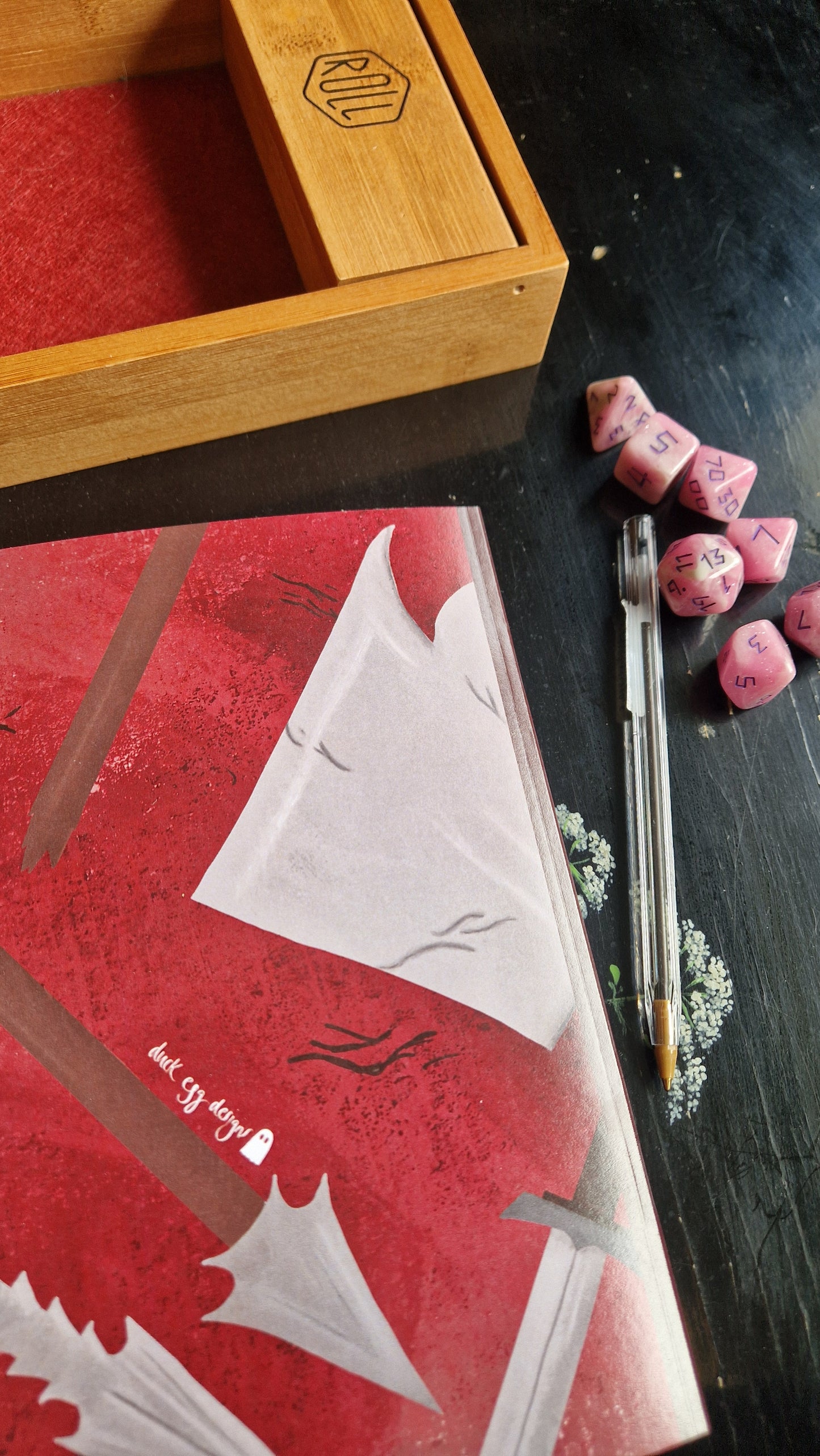 A tabletop RPG setup featuring a wooden dice tray, pink polyhedral dice, a clear pen, and campaign notes in the Duck Egg Designs Reckless Attack Notebook with illustrated weapons and "dark city story" on the cover, all on a black surface.