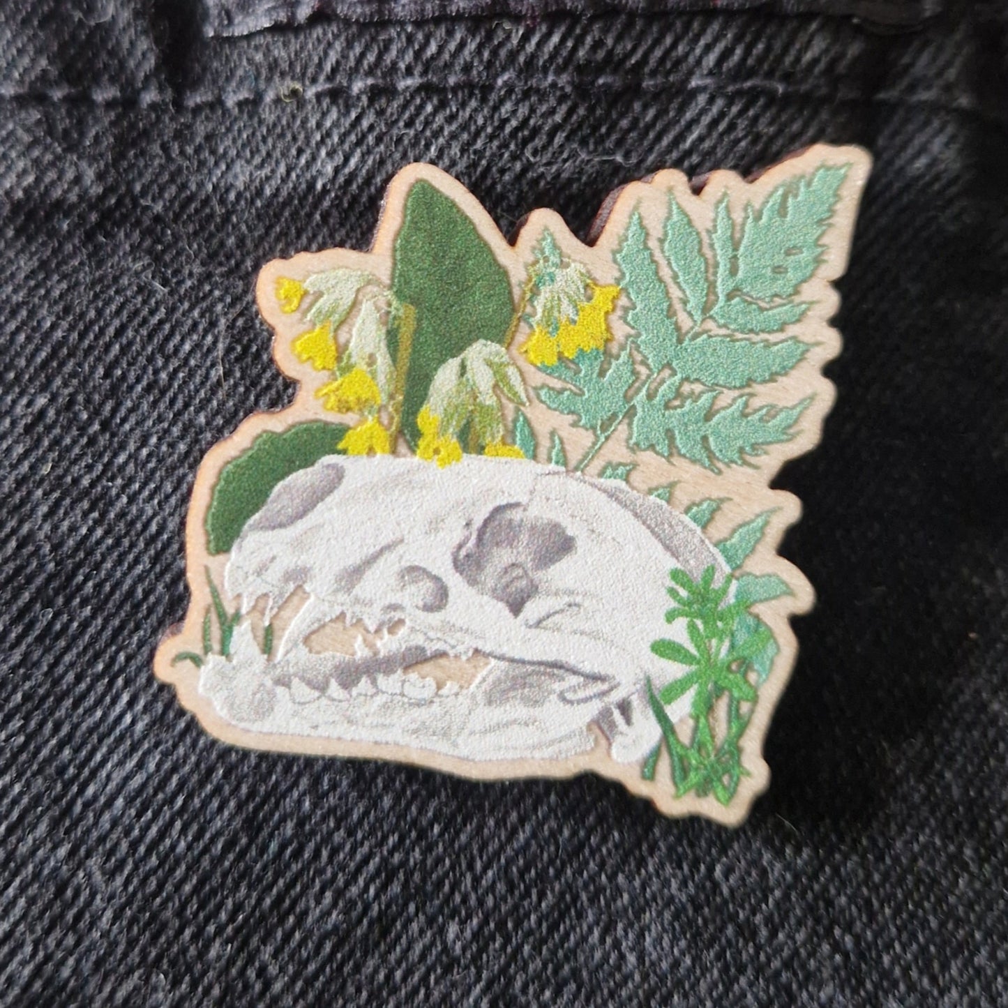Decorative pin badge with skull and leaves on dark grey denim fabric.