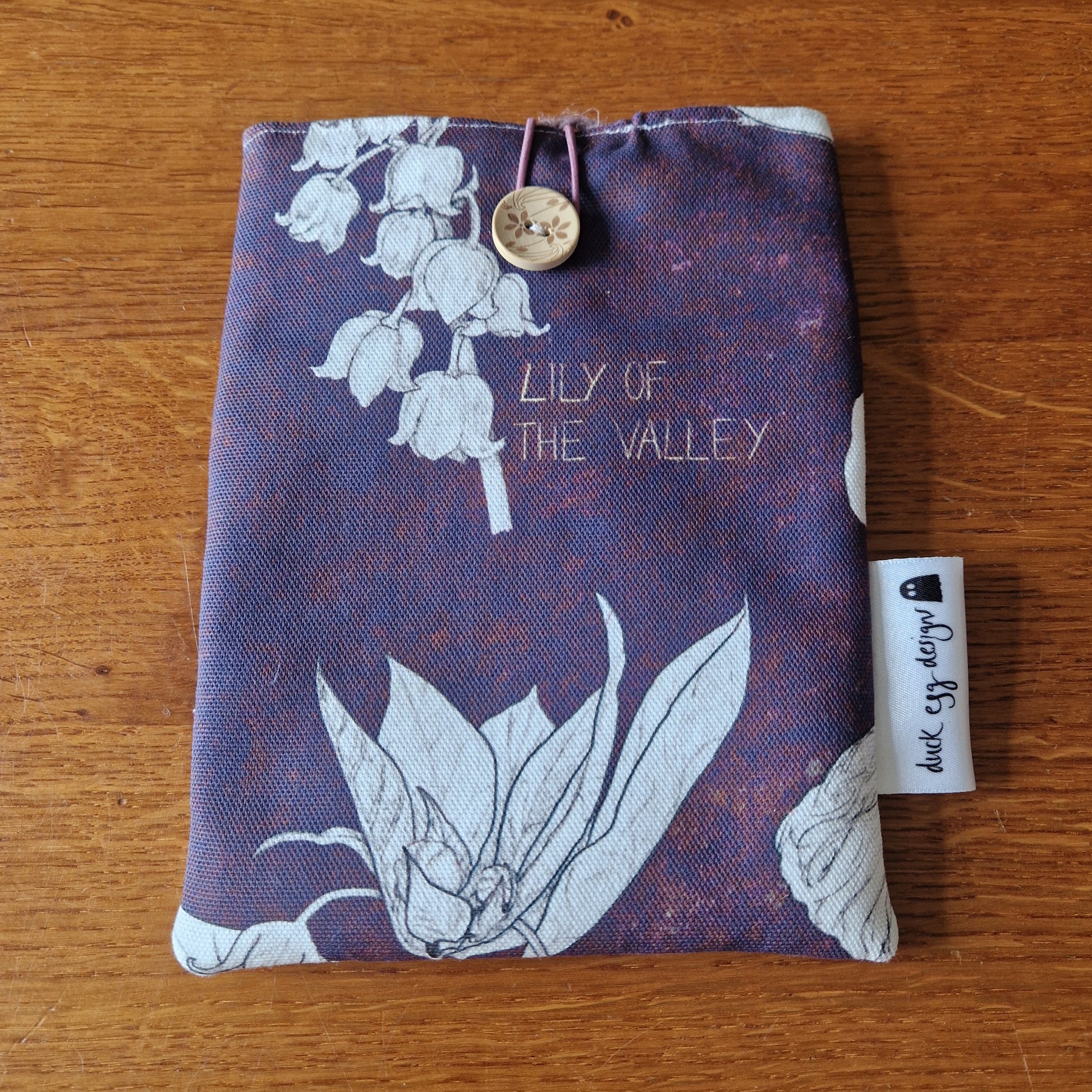 The Poison Plants Kindle Pouch by Duck Egg Designs features a handmade design with a Lily of the Valley print, button and loop closure, and a small branded fabric tag on the side. It is pictured resting on a wooden surface.
