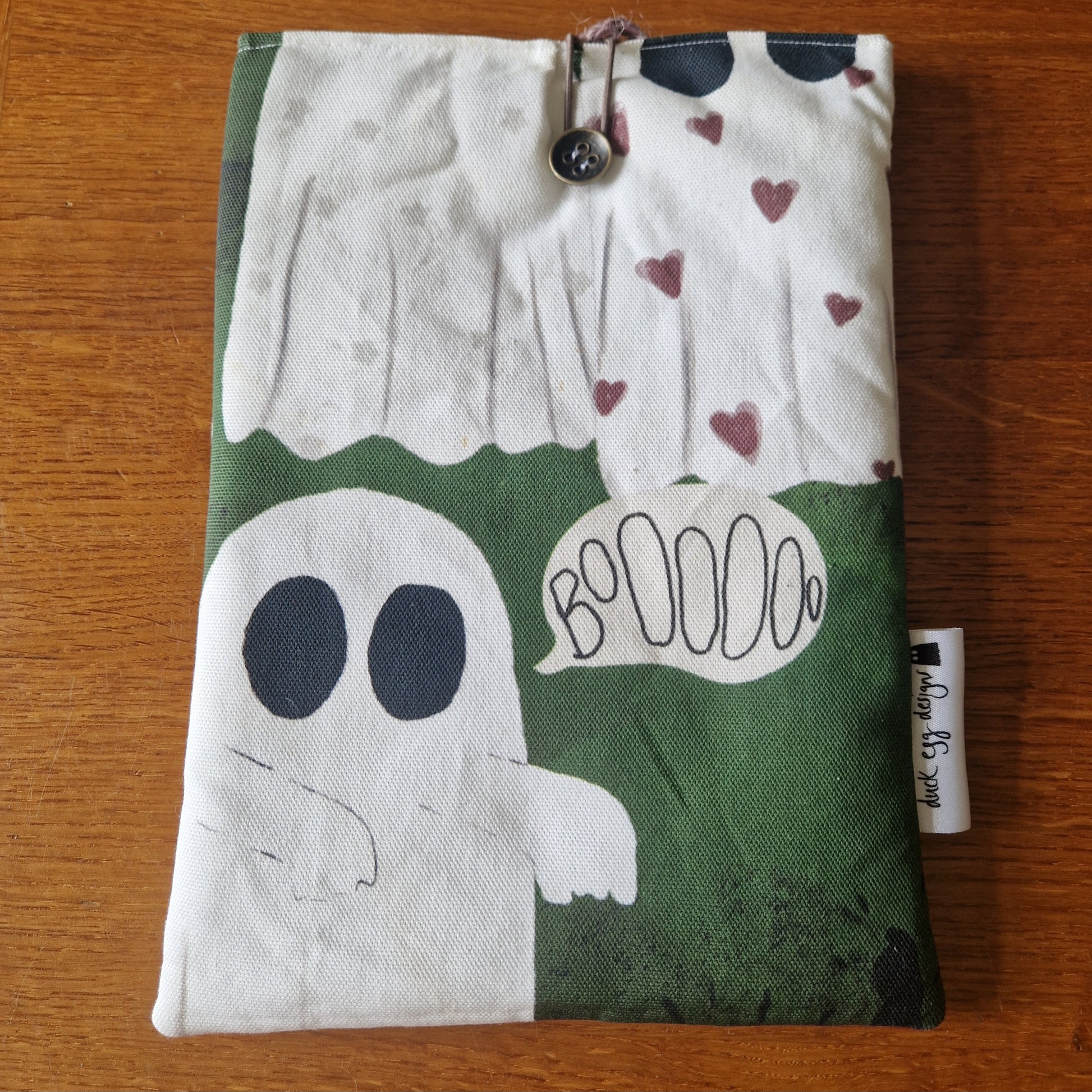 The A Haunting Of Ghosts Book Sleeve by Duck Egg Designs is a handmade pouch made from recycled fabric, decorated with cartoon ghosts and details like a button closure, side tag, and displayed on wood.
