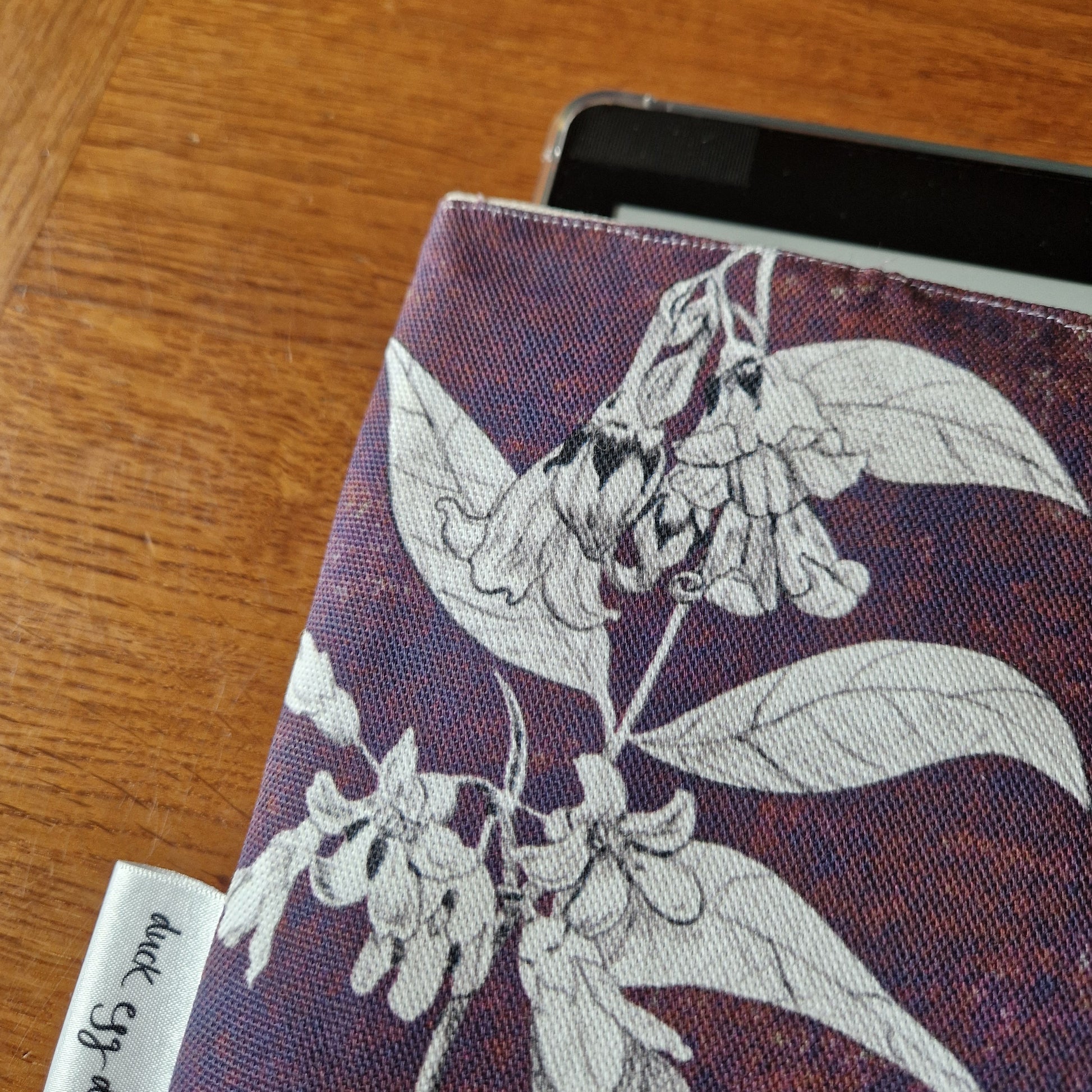 The Poison Plants Kindle Pouch by Duck Egg Designs features white floral patterns on a dark, handmade sleeve with a small fabric tag, shown resting on a wooden surface.