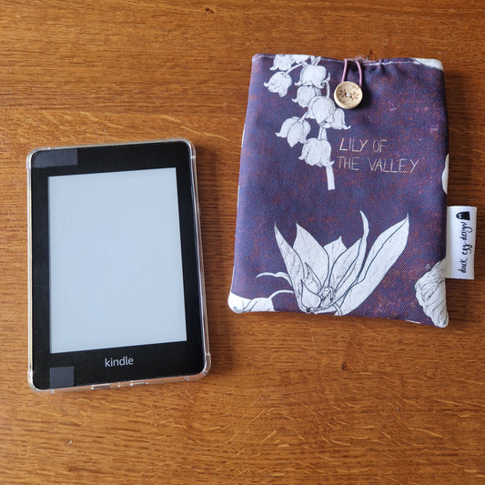 A Kindle e-reader with a black case rests on a wooden surface beside the Duck Egg Designs Poison Plants Kindle Pouch—a purple fabric sleeve featuring white lily of the valley flowers and a button.