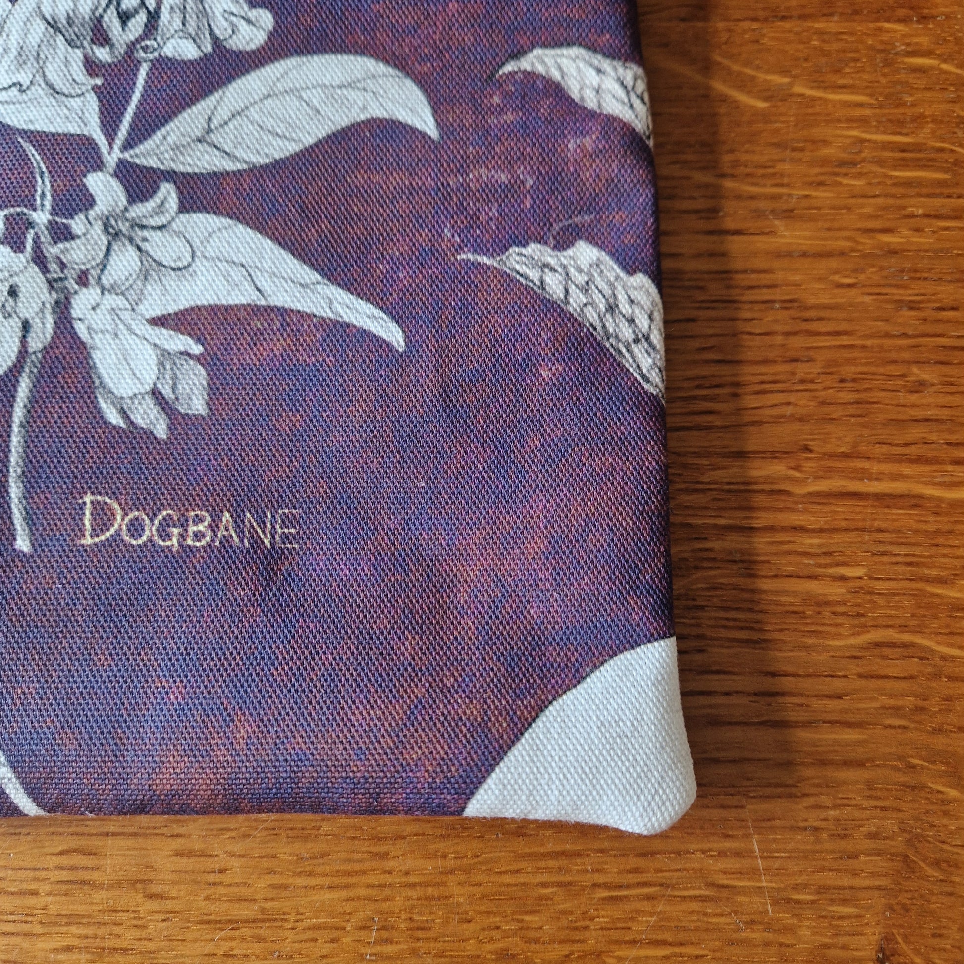 A close-up of the Poison Plants Kindle Pouch by Duck Egg Designs, featuring purple and maroon fabric, white botanical prints, and DOGBANE in gold text. This handmade case rests on a wooden surface.