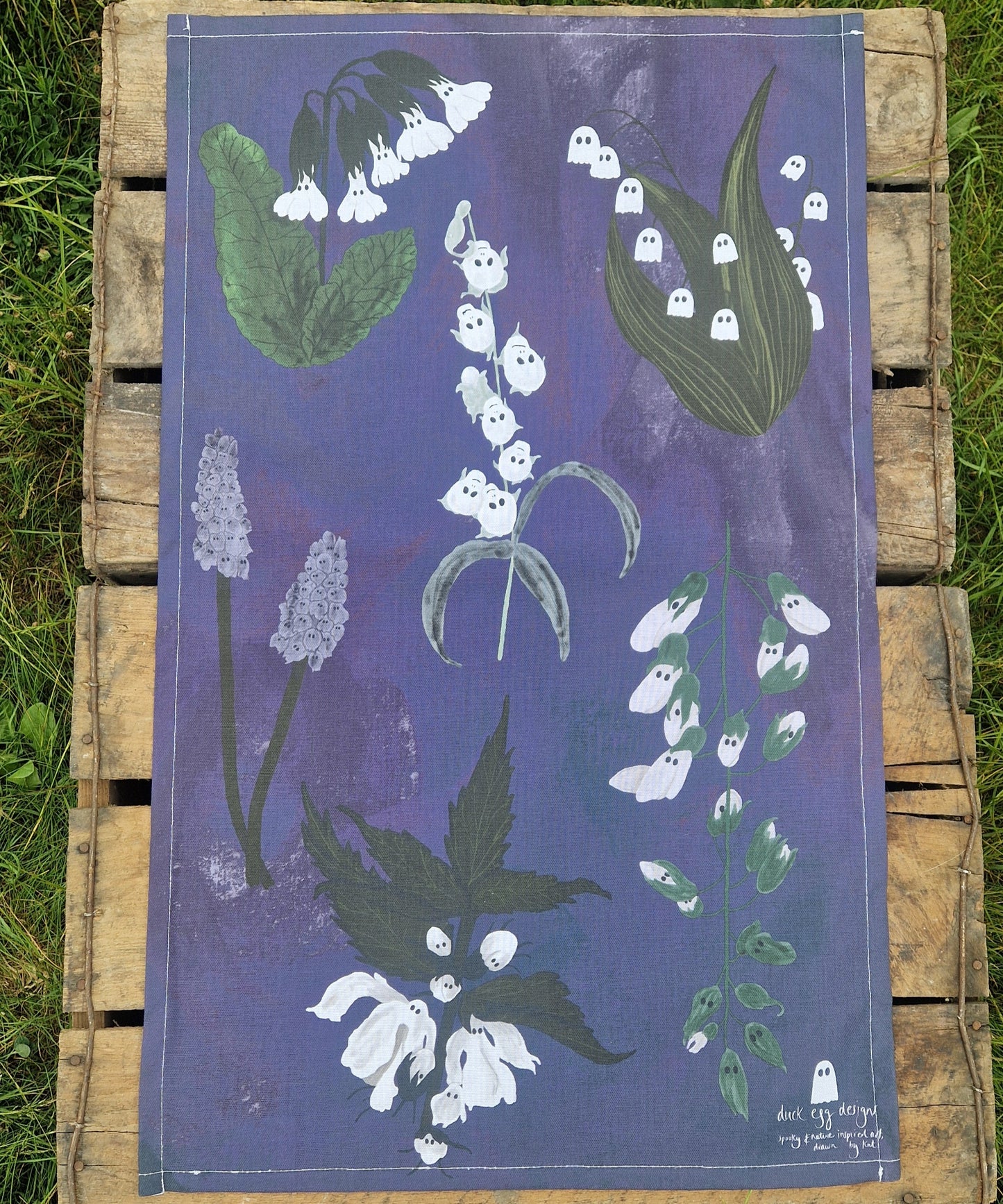 The Spooky Flowers Tea Towel by Duck Egg Designs features botanical white flowers with ghost faces on a purple background, displayed on wood with grass around the edges.