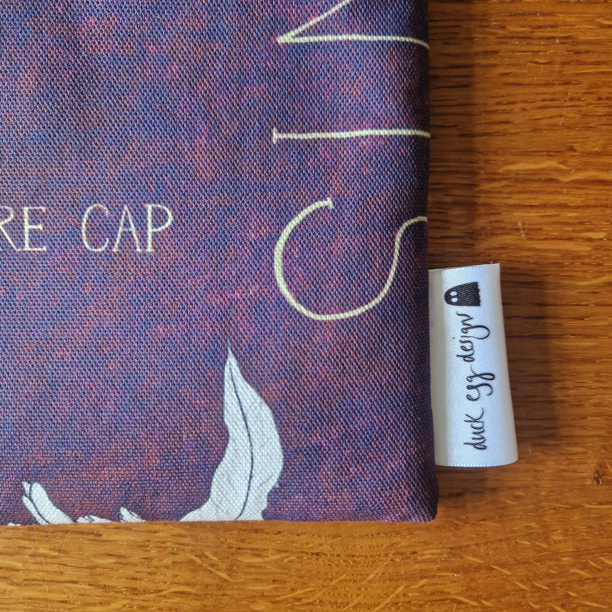 A close-up of the Poison Plants Book Sleeve by Duck Egg Designs shows purple upcycled fabric with white and yellow print, part of the words RE CAP, and a duck egg design label on a wooden surface.