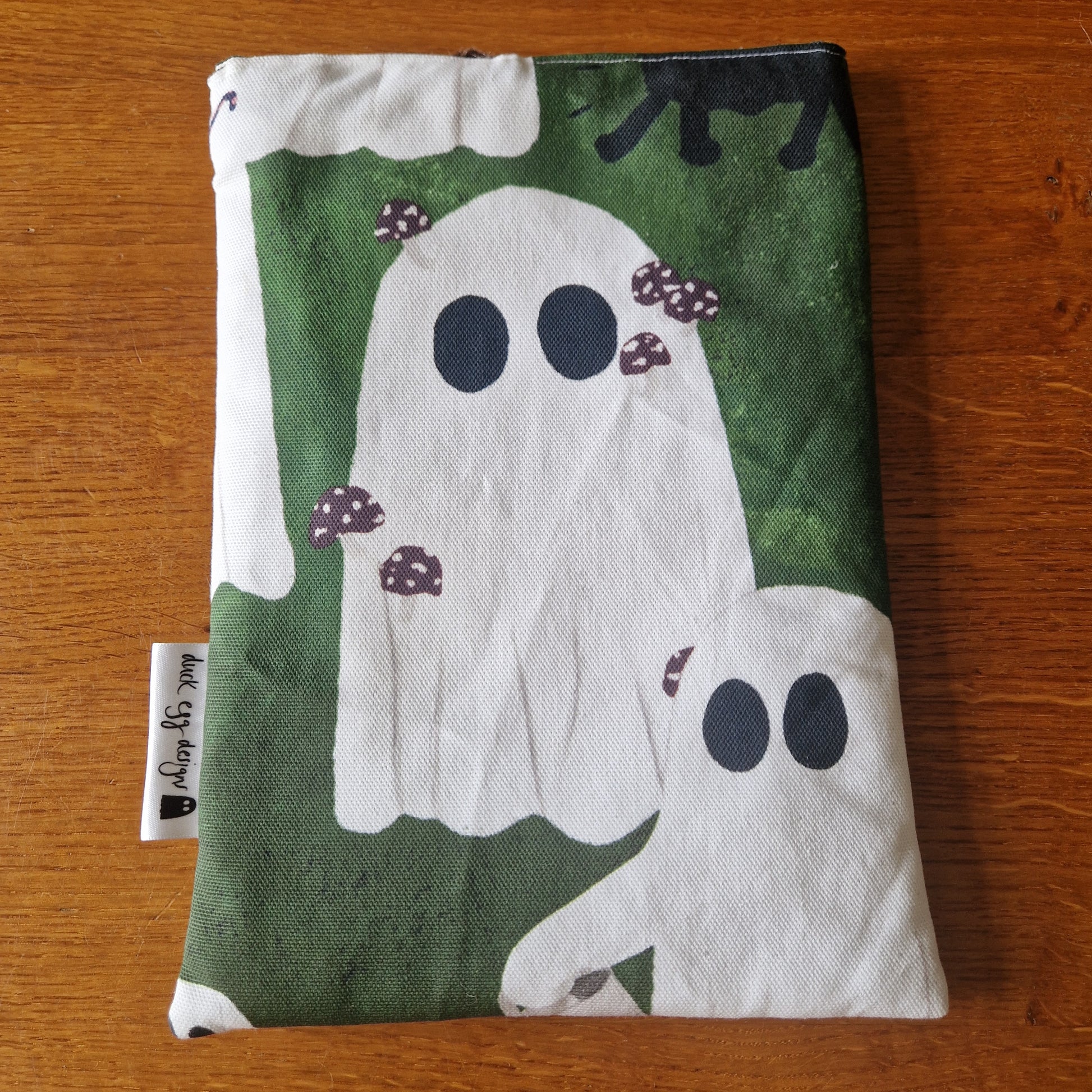 The A Haunting Of Ghosts Book Sleeve by Duck Egg Designs is a handmade fabric pouch with a green background, white ghosts with black eyes and tiny brown mushrooms, plus a small side tag—perfect for adding quirky charm to your books.