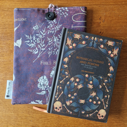 A Poison Plants Book Sleeve by Duck Egg Designs, crafted from upcycled fabric with botanical illustrations and plant names, sits on a wooden surface beneath the Botanical Curses and Poisons book.