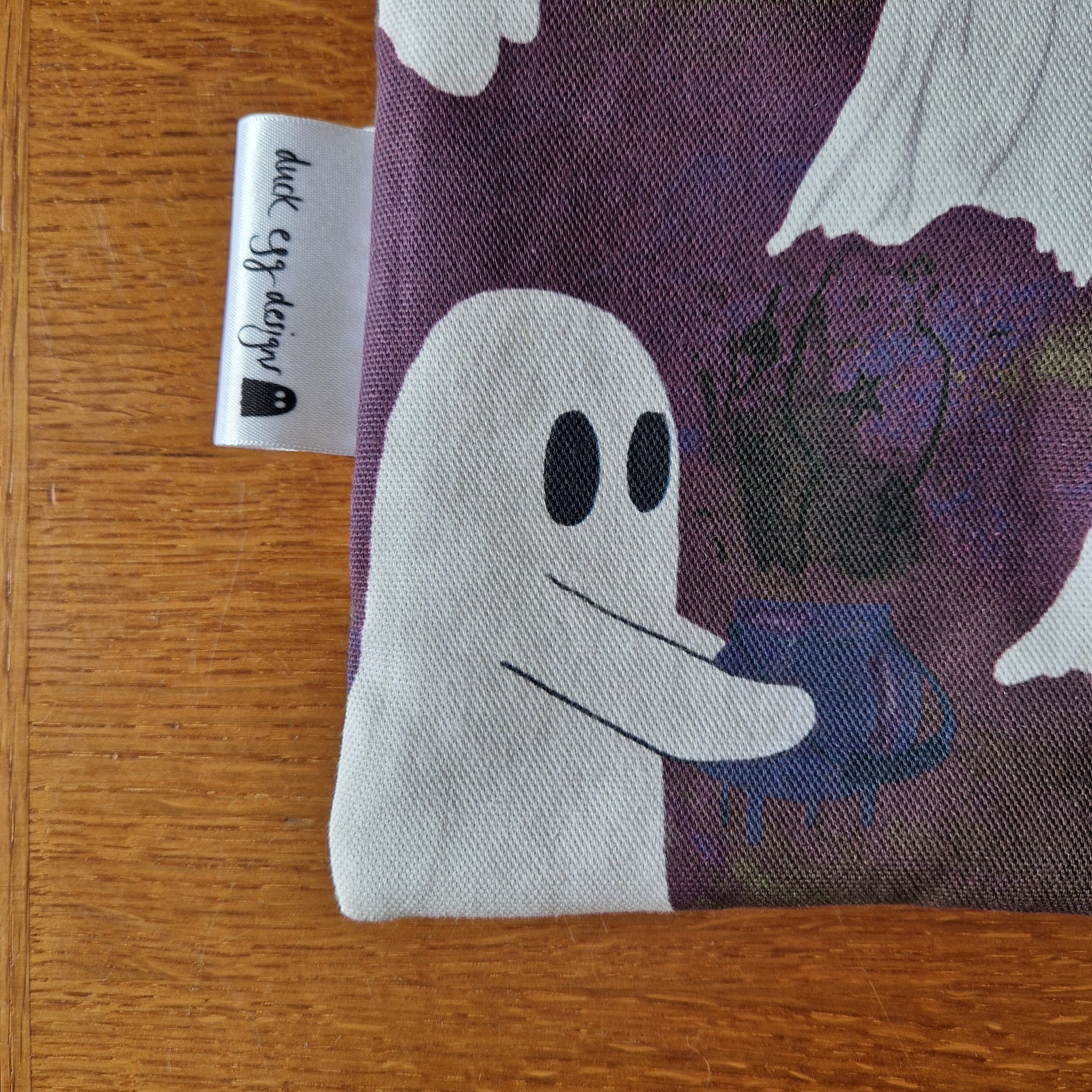 The A Fright Of Ghosts Book Sleeve by Duck Egg Designs is made from upcycled fabric and features a white ghost with a blue bowl on purple, plus a branded tag—perfect as a reusable book cover or book sleeve.