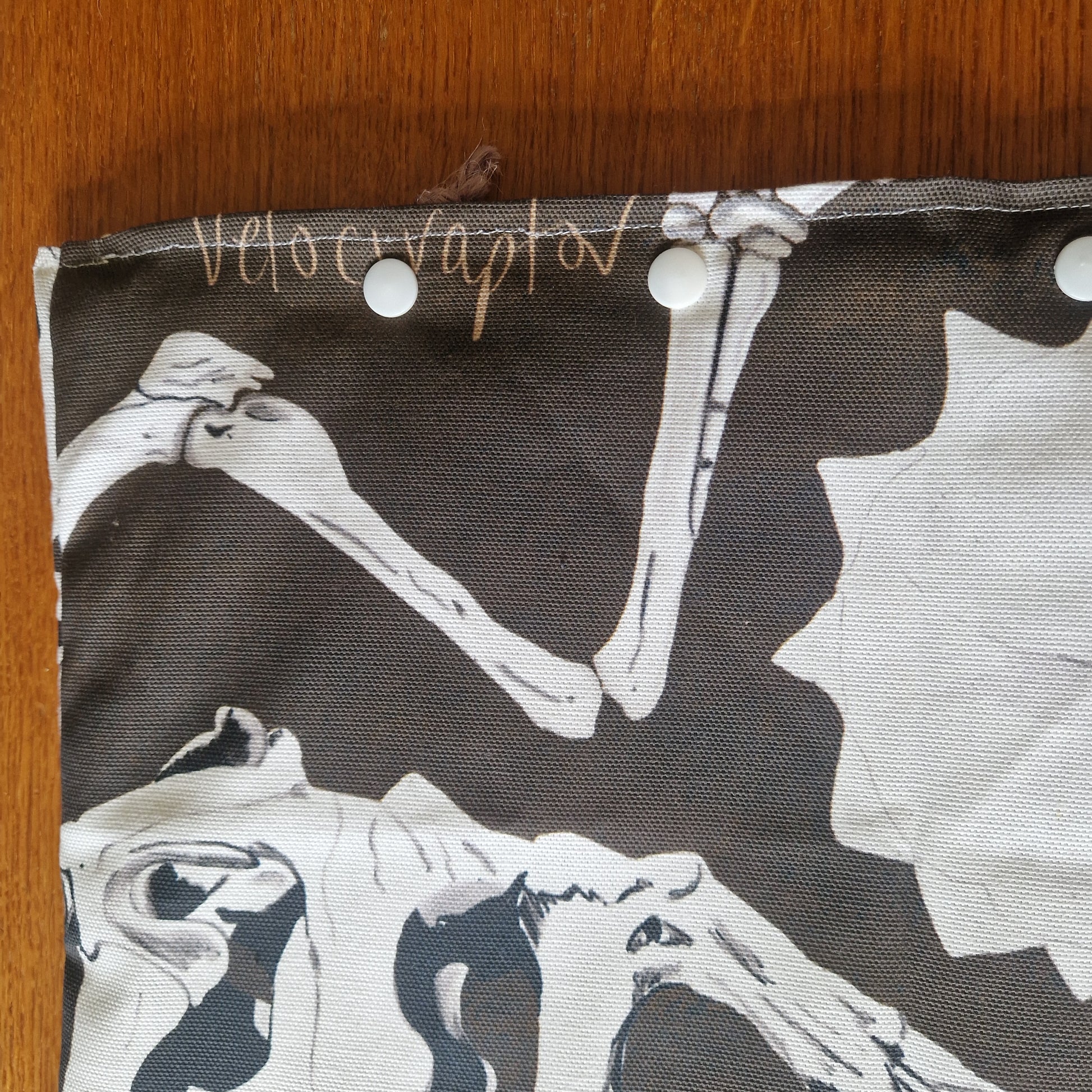 The Dinosaur Bones Small Laptop/Tablet Sleeve by Duck Egg Designs is handmade from recycled fabric, with white dinosaur bones on dark fabric, Velociraptor handwritten on top, and white snap buttons along the edge.