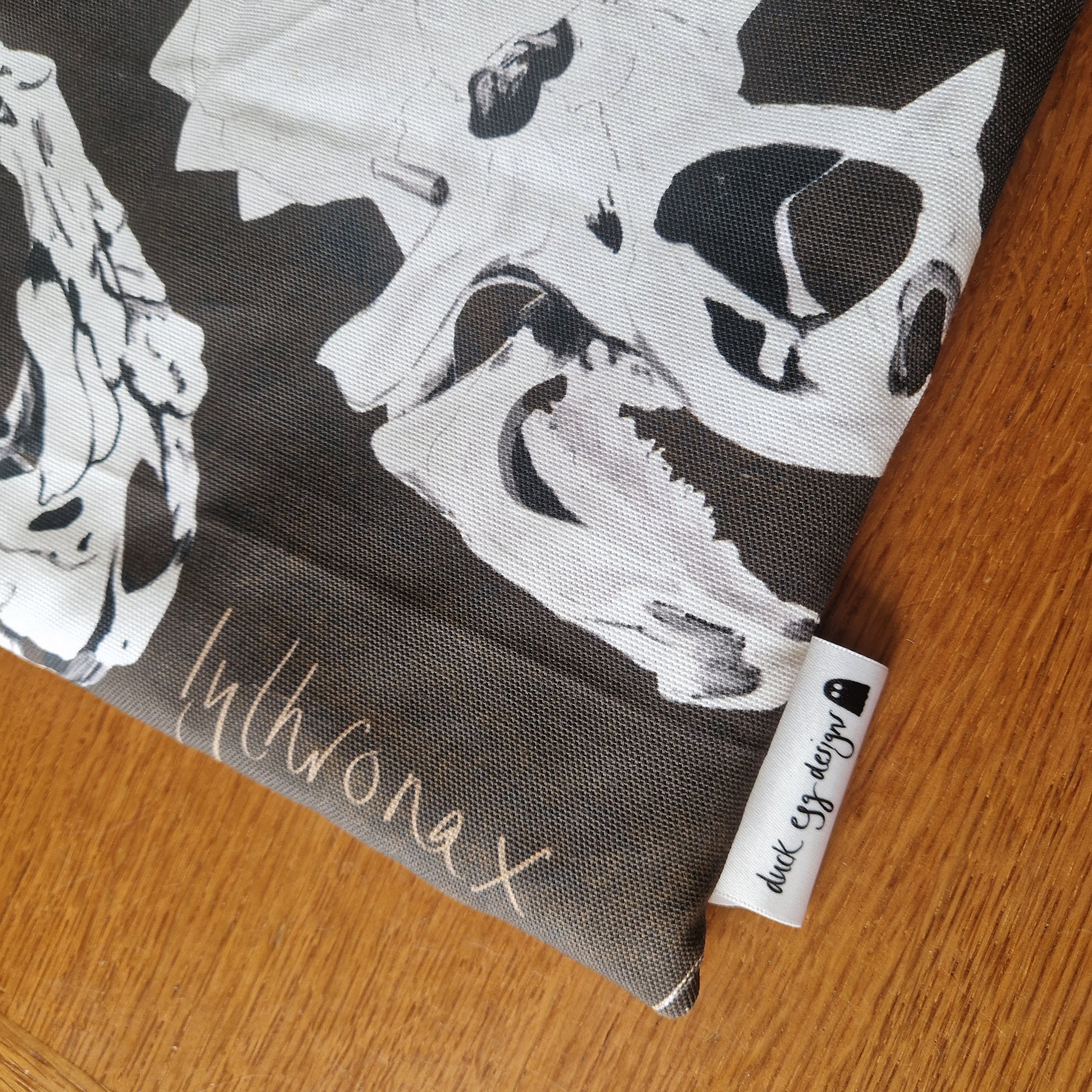 The Dinosaur Bones Small Laptop/Tablet Sleeve by Duck Egg Designs is handmade from recycled fabric, with white dinosaur skulls on dark material, Duck Egg Designs tag, a duck icon, and displayed on a wooden surface.