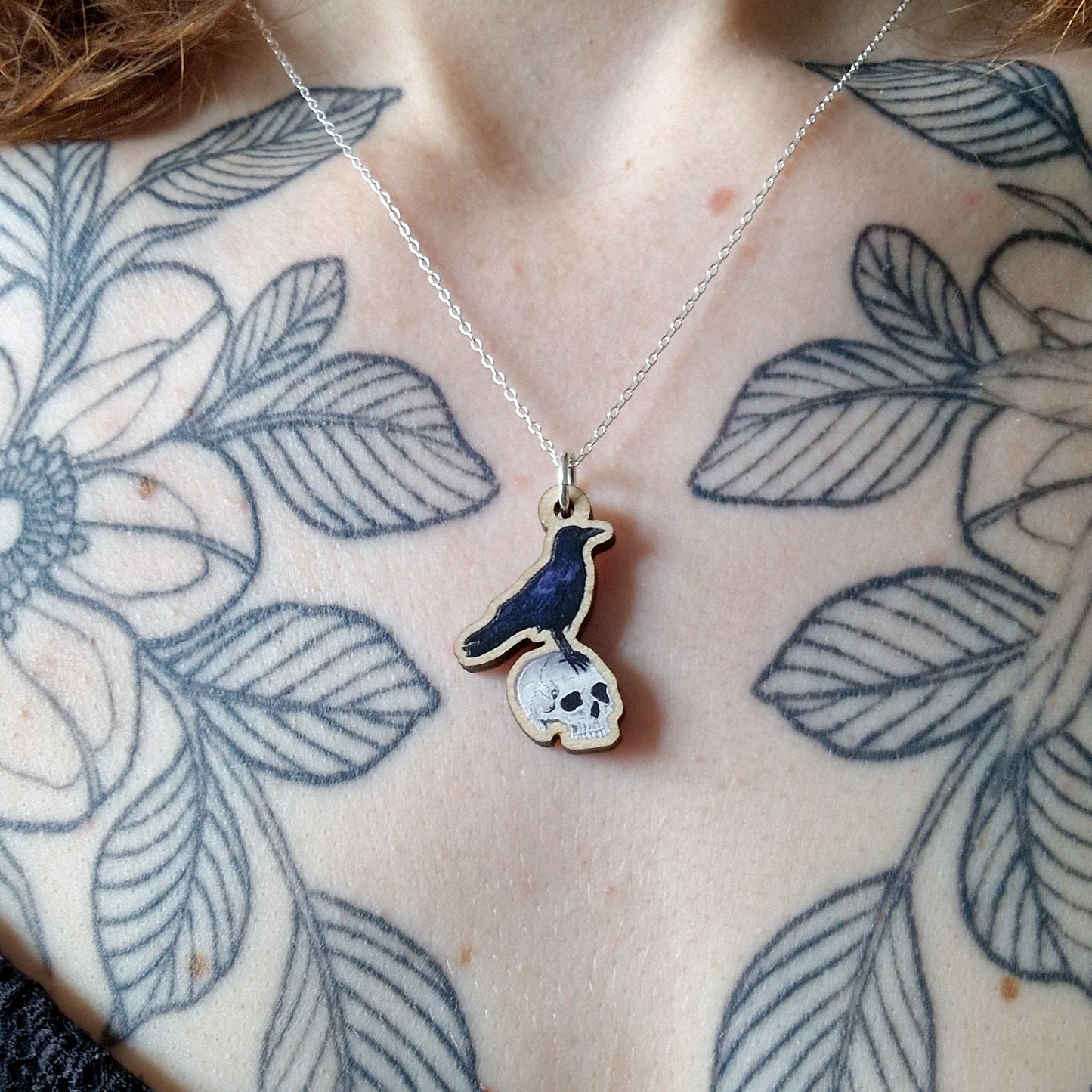 Necklace with a pendant featuring a bird and skull design, worn by a person with leaf tattoos on their shoulders and chest.
