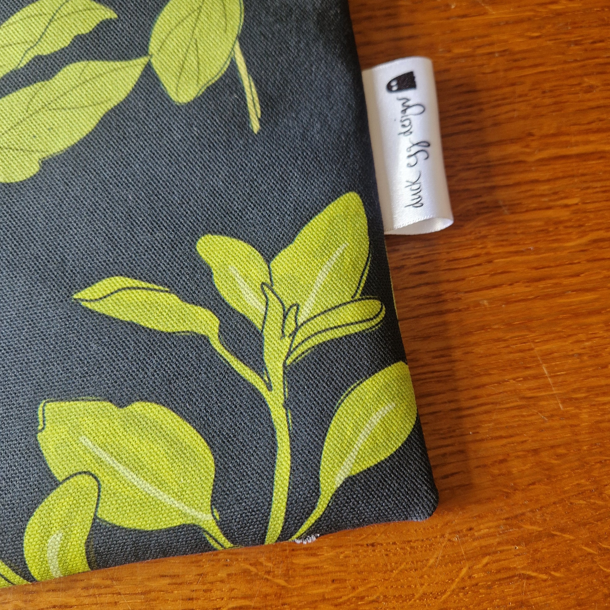 The Common Herbs Kindle Pouch by Duck Egg Designs, featuring a green leaf pattern on a dark background, rests on a wooden surface. A white tag with the brand name is visible on the right edge of this unique Kindle case.