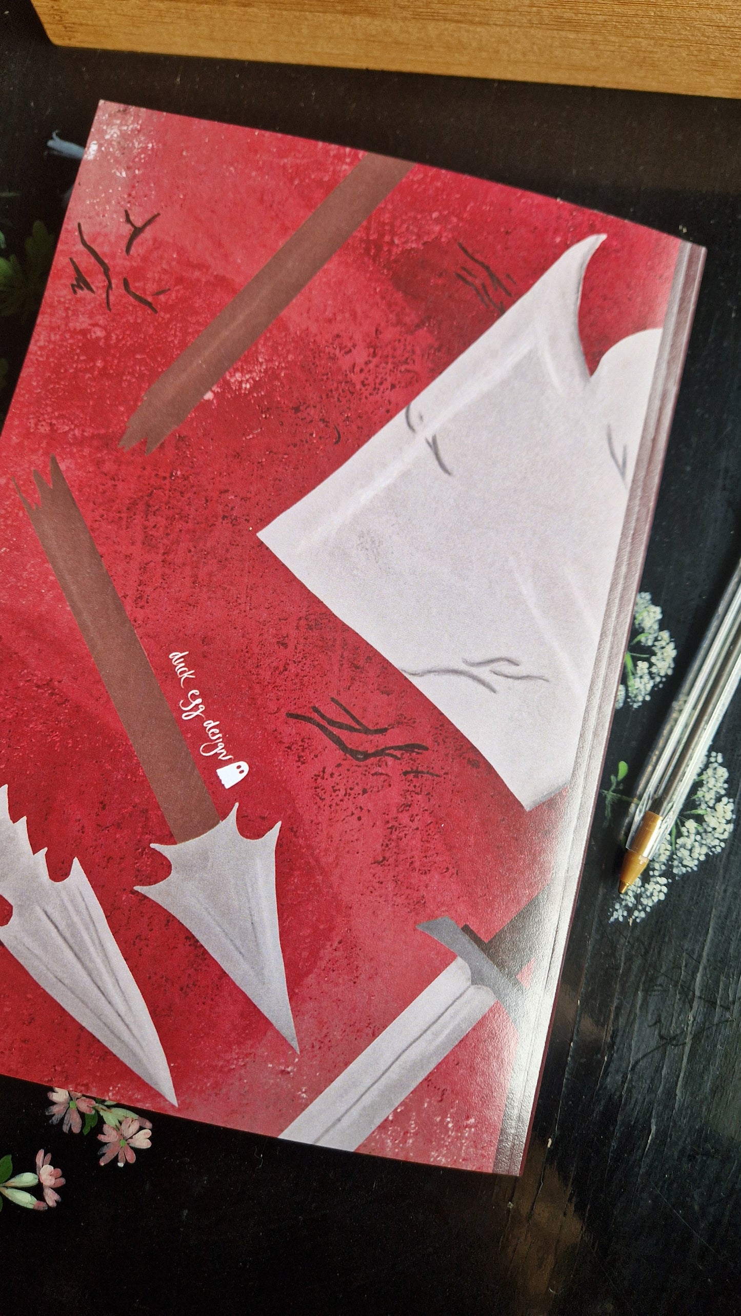 The Reckless Attack Notebook by Duck Egg Designs features a red cover with illustrated swords, white torn cloth, and scratch marks. "The Ghost Story" and a small ghost are printed on recycled paper. A pen and flowers rest nearby on the dark surface.