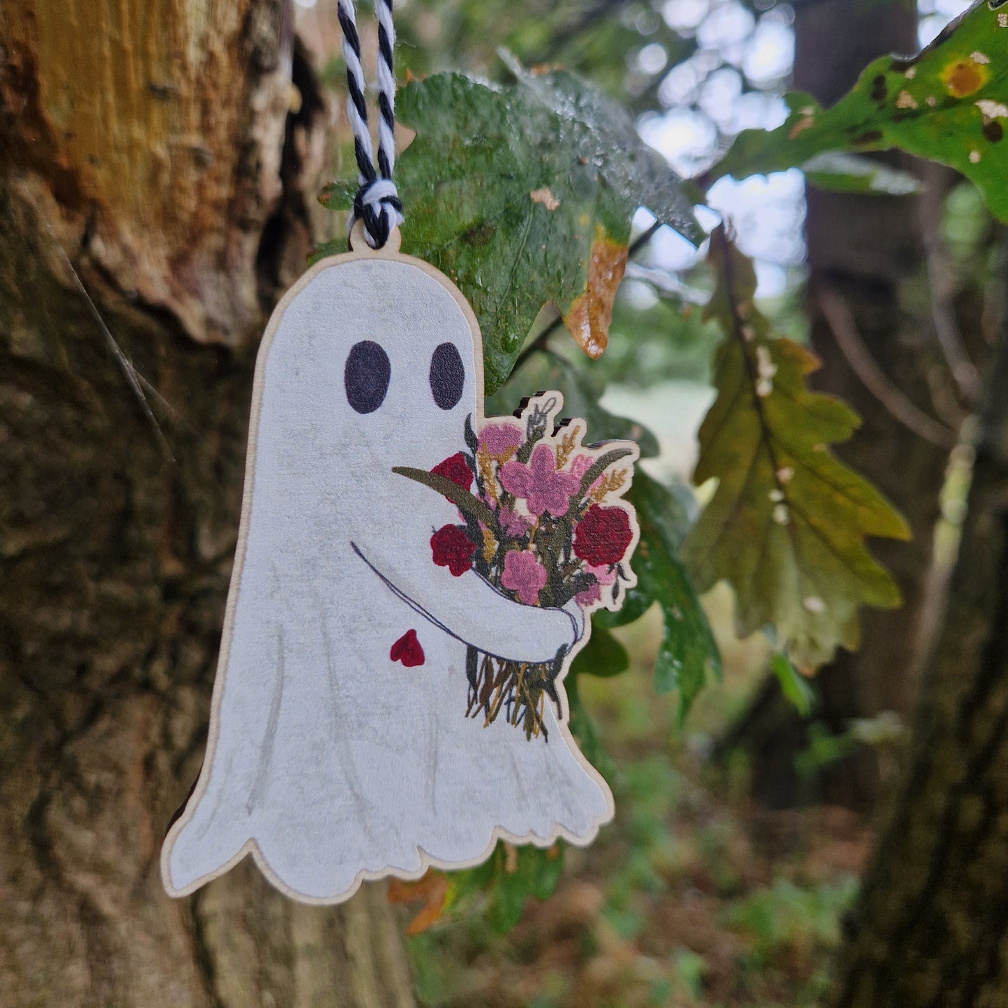 The Flower Ghost Hanging Wooden Decoration by Duck Egg Designs features a whimsical illustrated ghost on wood, suspended by a black and white string from a tree branch with green leaves and blurred foliage in the background.