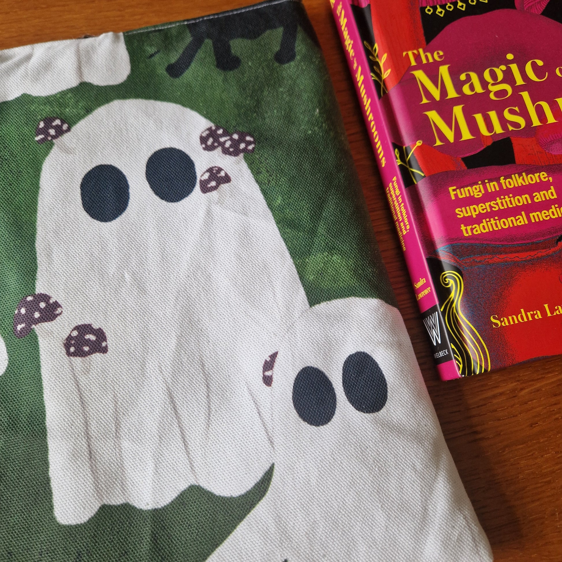 The A Haunting Of Ghosts Book Sleeve by Duck Egg Designs, handmade from recycled fabric with ghosts featuring mushroom spots, rests next to The Magic of Mushrooms by Sandra Lawrence on a wooden surface.
