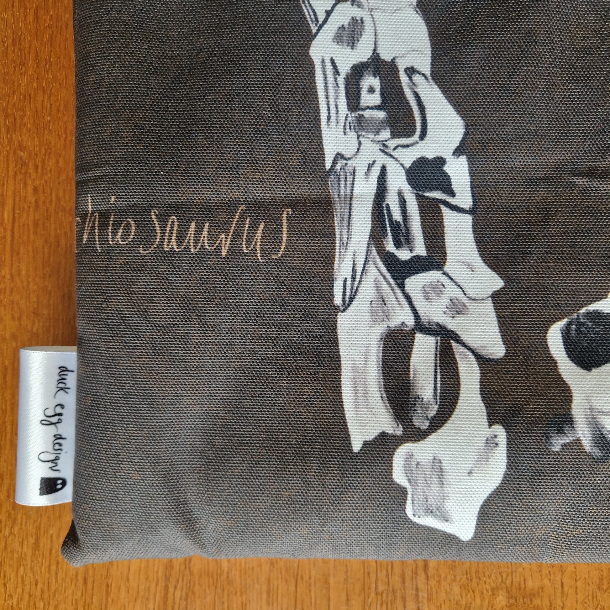 A close-up of dark, recycled fabric printed with white dinosaur bones and dinosaurs in script, plus a Duck Egg Designs tag, highlights the handmade Dinosaur Bones Small Laptop/Tablet Sleeve.