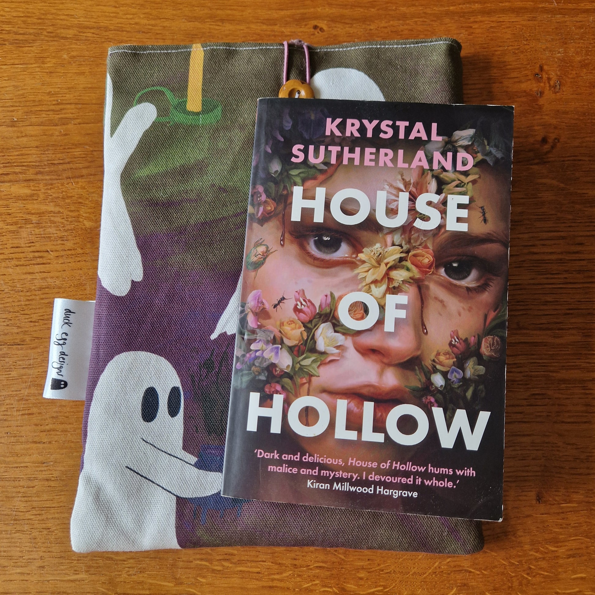 A copy of House of Hollow by Krystal Sutherland sits on Duck Egg Designs’ A Fright Of Ghosts Book Sleeve, made from upcycled fabric with a ghost print, protecting the striking cover as it rests on a wooden surface.
