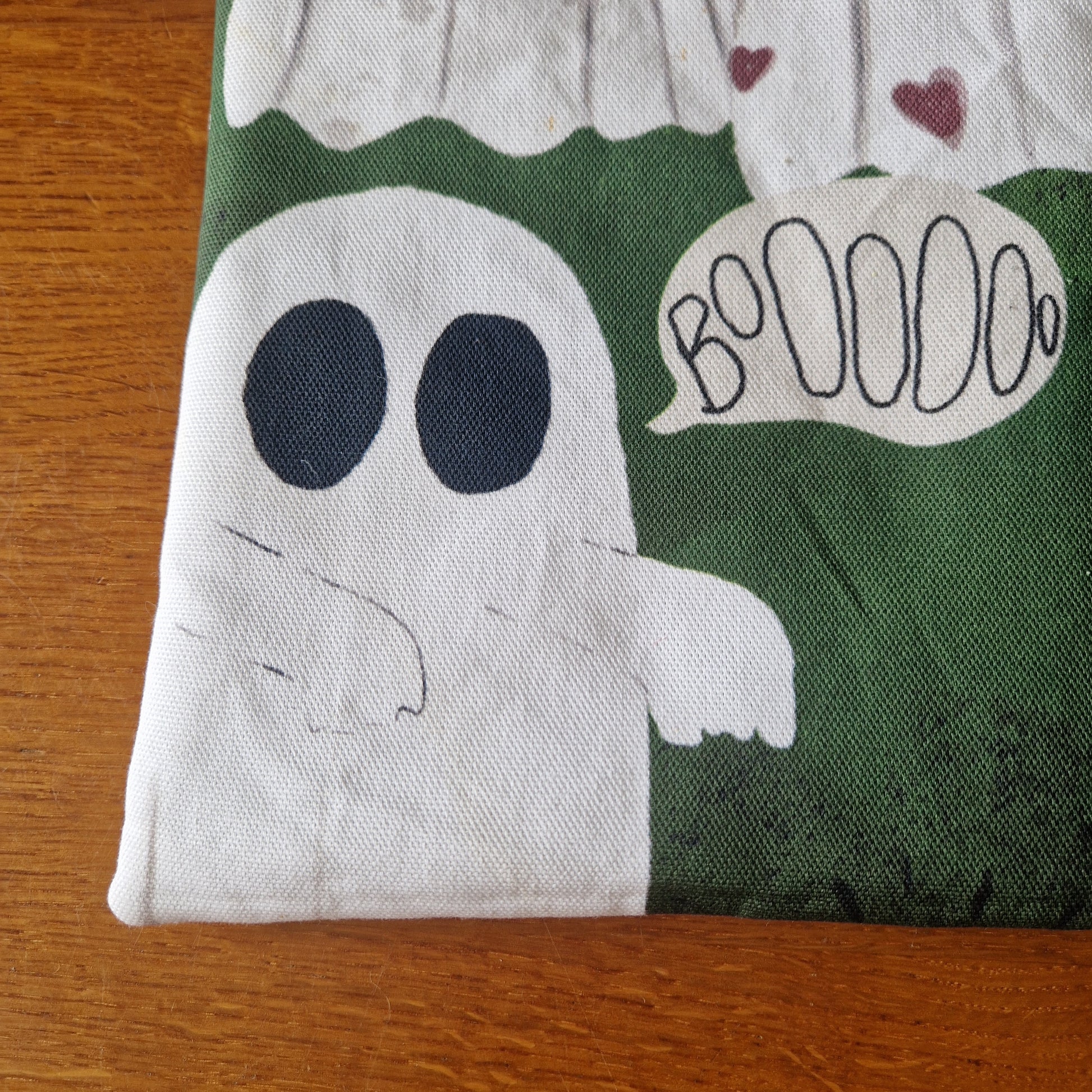 The A Haunting Of Ghosts Book Sleeve by Duck Egg Designs is handmade from recycled fabric, featuring a cute white ghost with black eyes and a Boooo bubble on a green background, shown on a wooden surface.