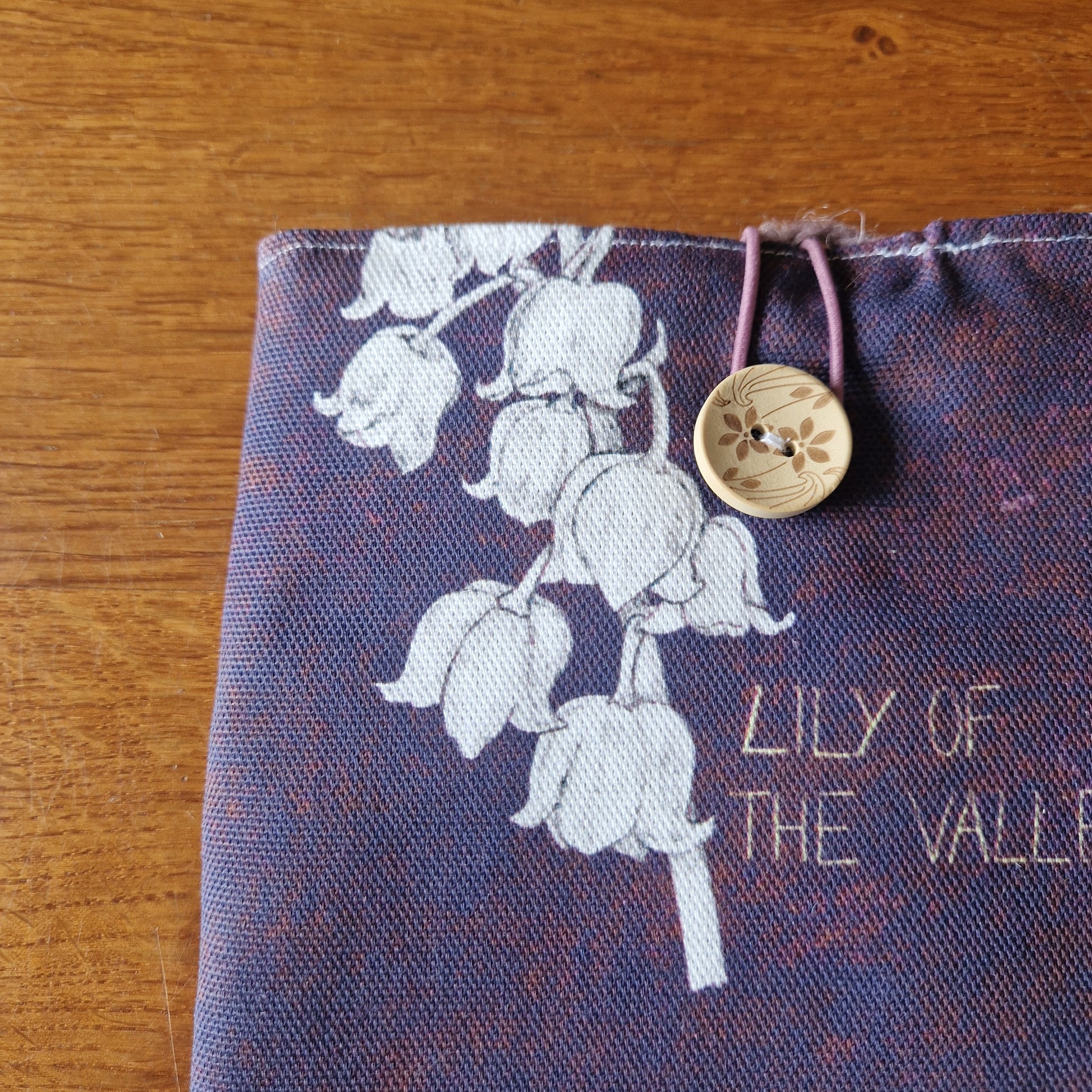 The Poison Plants Kindle Pouch by Duck Egg Designs is a purple fabric sleeve featuring white lily of the valley print, gold LILY OF THE VALLEY embroidery, and a beige floral button on a pink elastic loop, shown on a wooden surface.