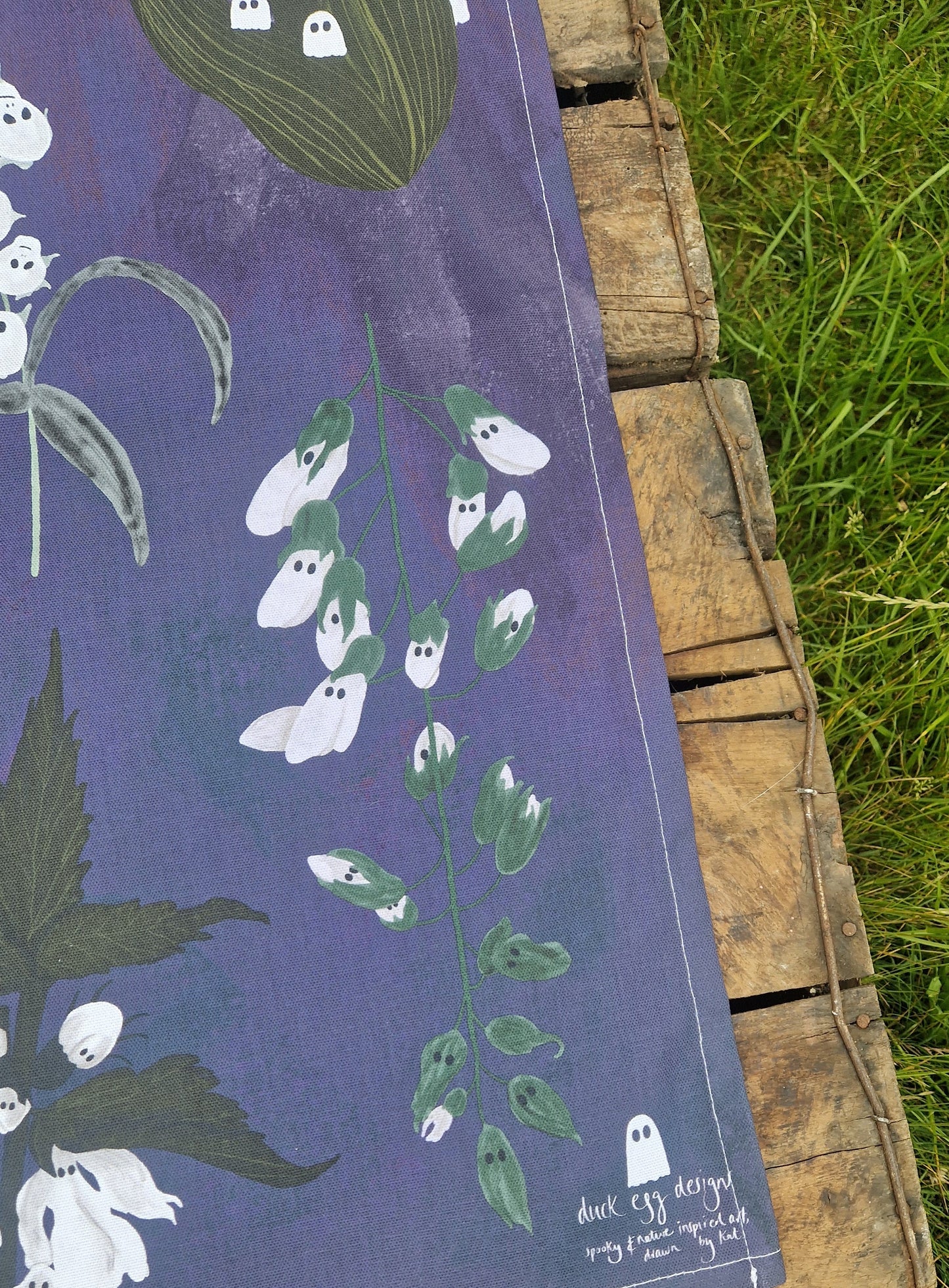The Spooky Flowers Tea Towel by Duck Egg Designs features white flowers with ghost-like faces on a purple background, draped over a wooden pallet on grass and signed in the corner.