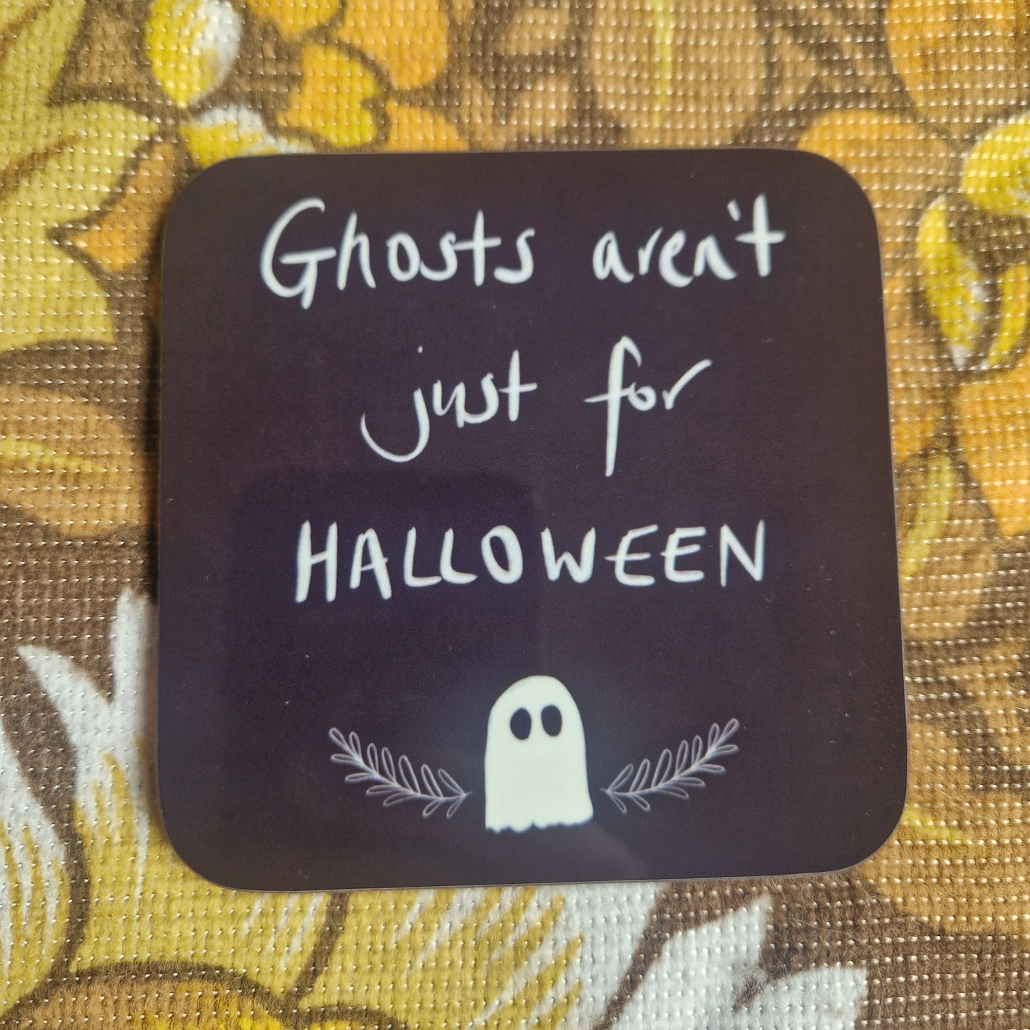 The Ghosts Aren't Just For Halloween Coaster by Duck Egg Designs is a square UK-made coaster with white handwritten text above a simple ghost and leaves, shown on a yellow floral-patterned background.