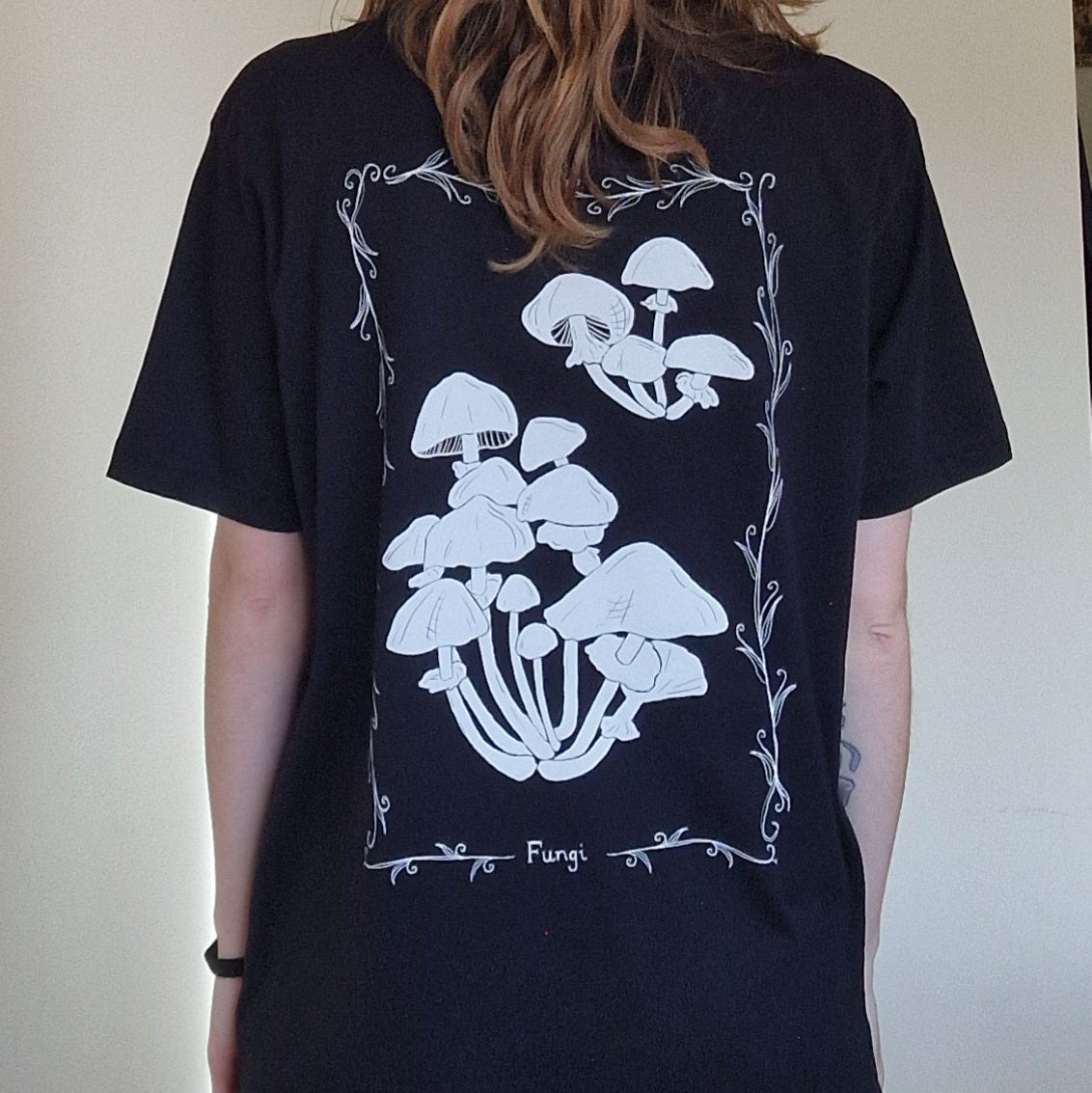 A person wears the Duck Egg Designs Black Fungi Tshirt With Back And Front Print, featuring white mushroom illustrations framed by decorative lines and “Fungi” at the bottom. The shirt is shown from the back.