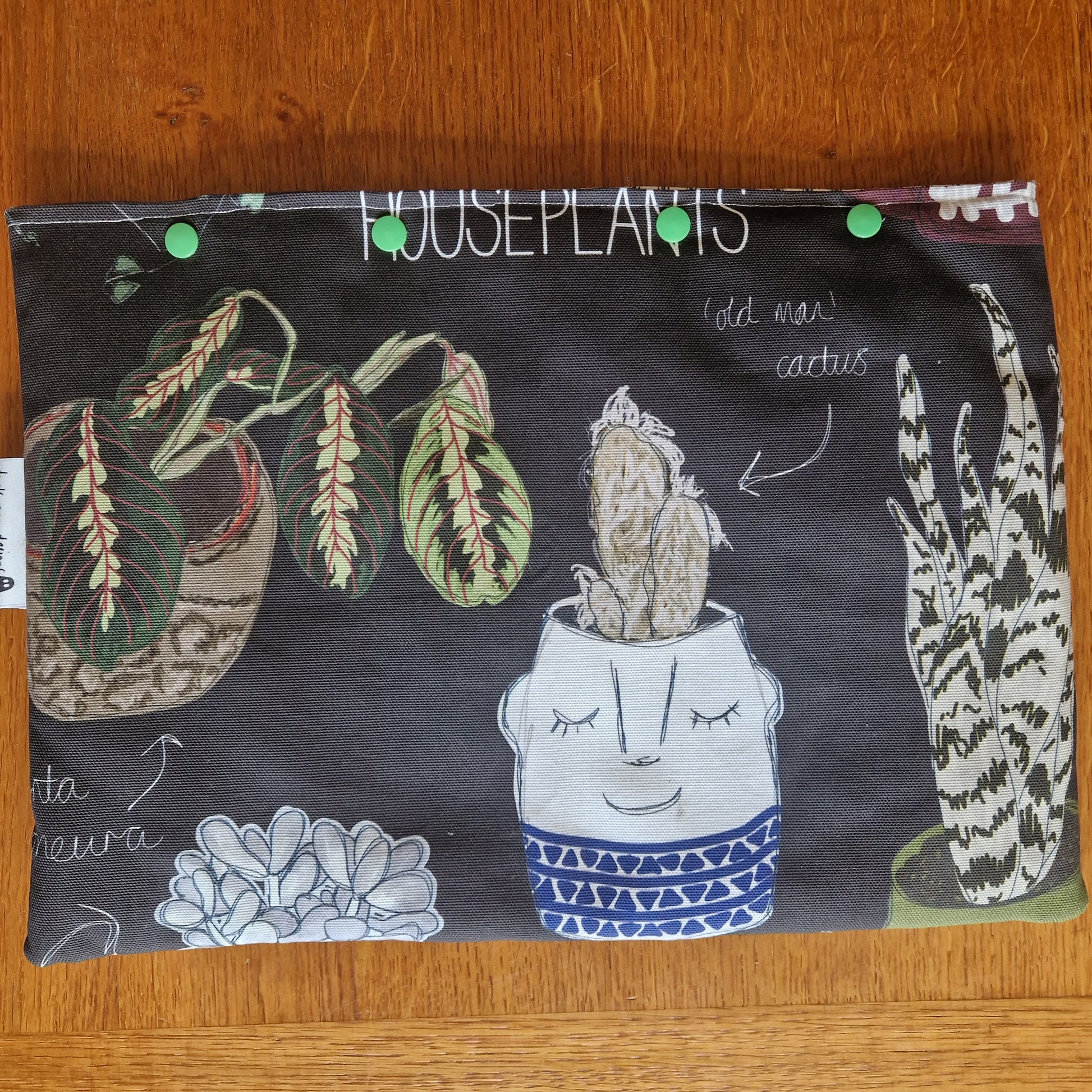 The Common Houseplants Small Laptop/Tablet Sleeve by Duck Egg Designs is handmade from recycled fabric. It features houseplant illustrations—one in a smiling-face pot—on a dark background with white handwritten plant names and green snaps.