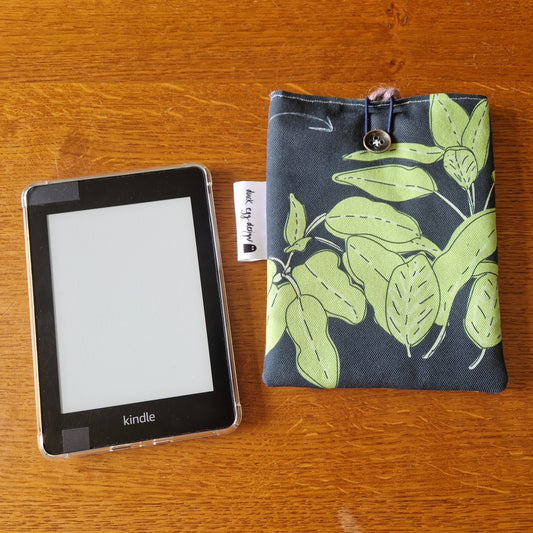 A black Kindle e-reader rests on a wooden surface beside the Duck Egg Designs Common Herbs Kindle Pouch—a fabric sleeve with green leaf patterns, button closure, and white tag—offering stylish protection.