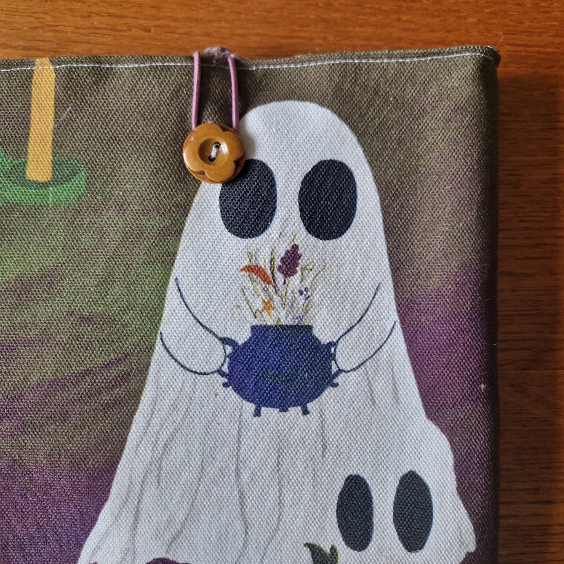 The A Fright Of Ghosts Book Sleeve by Duck Egg Designs features a ghost with a purple cauldron of dried flowers, printed on upcycled fabric. It has a pink elastic loop and wooden button, and is pictured on a wooden surface.