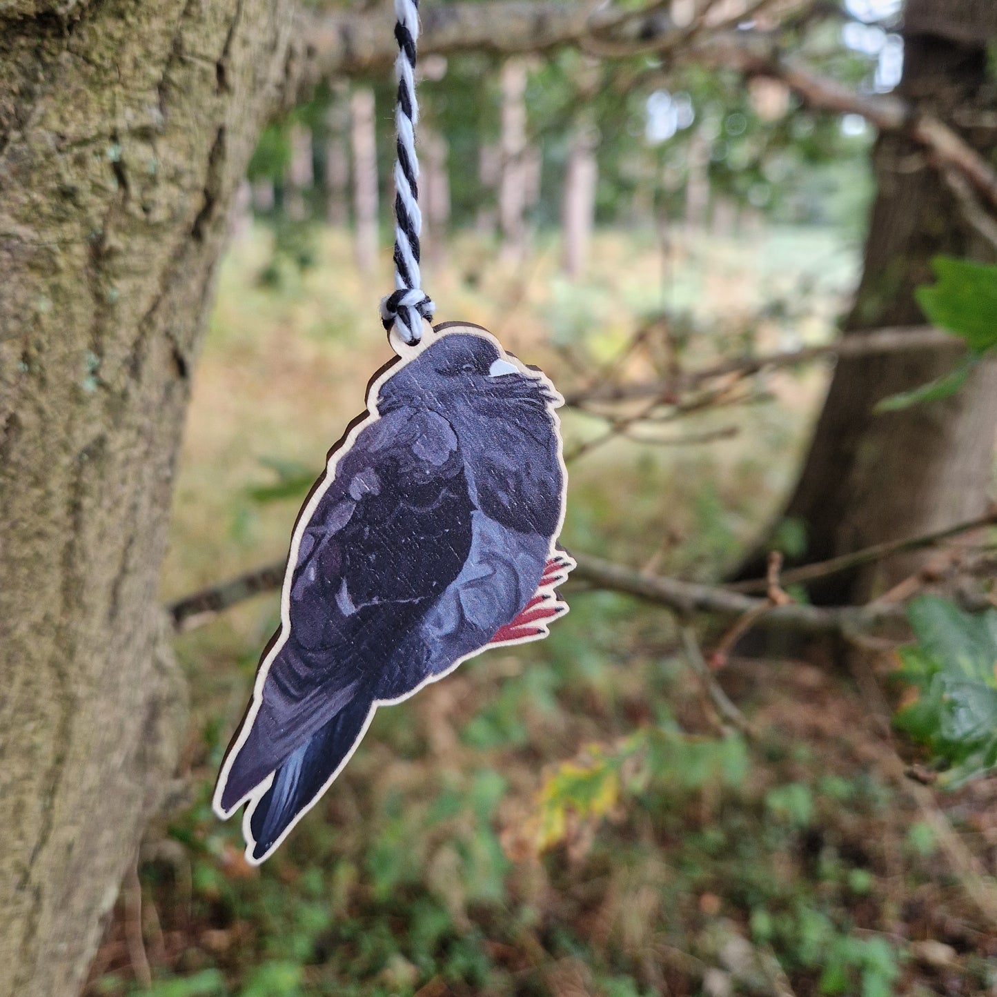 The Bob Chonky Pigeon Hanging Decoration by Duck Egg Designs hangs from a black-and-white string on a tree branch in the forest, with blurred trees and greenery in the background.