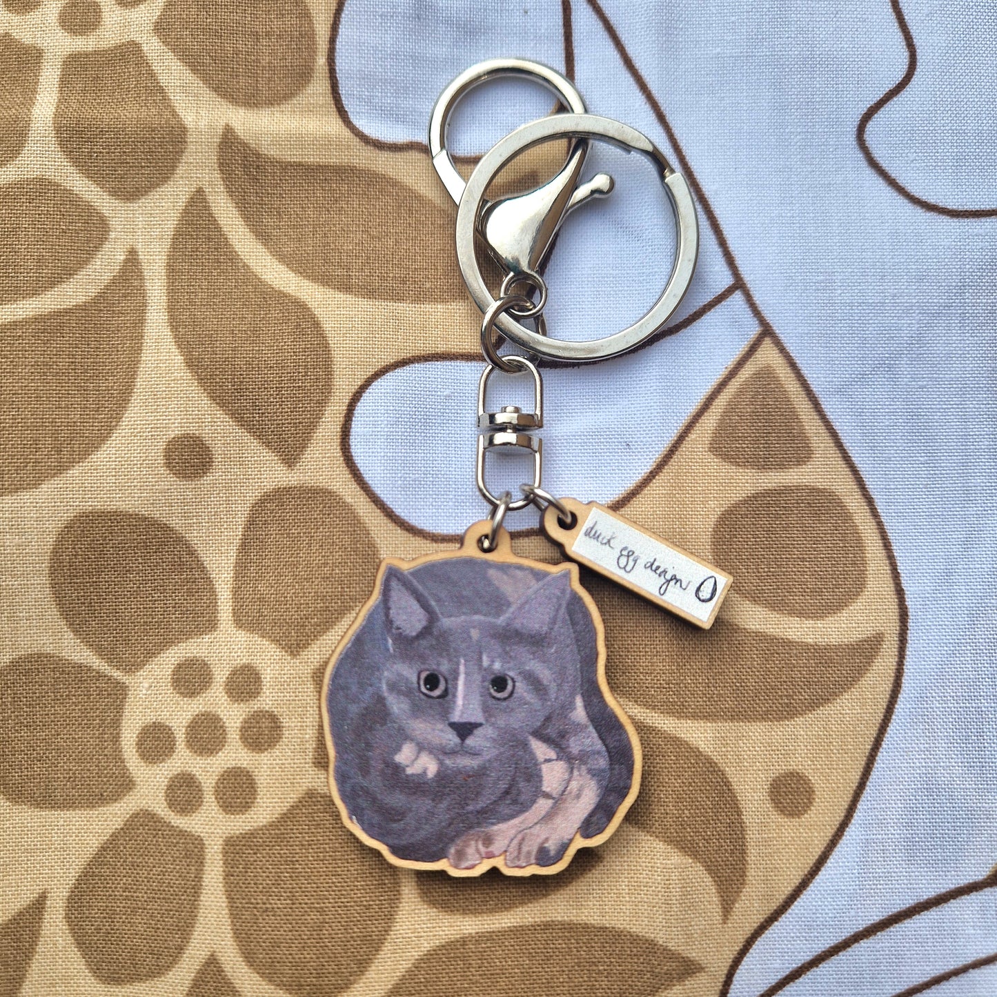 The Luna Grey Cat Keyring by Duck Egg Designs is a UK-made charm featuring an illustrated grey cat on sustainable wood and a metal tag reading "heck off, human," shown on brown and beige floral fabric.