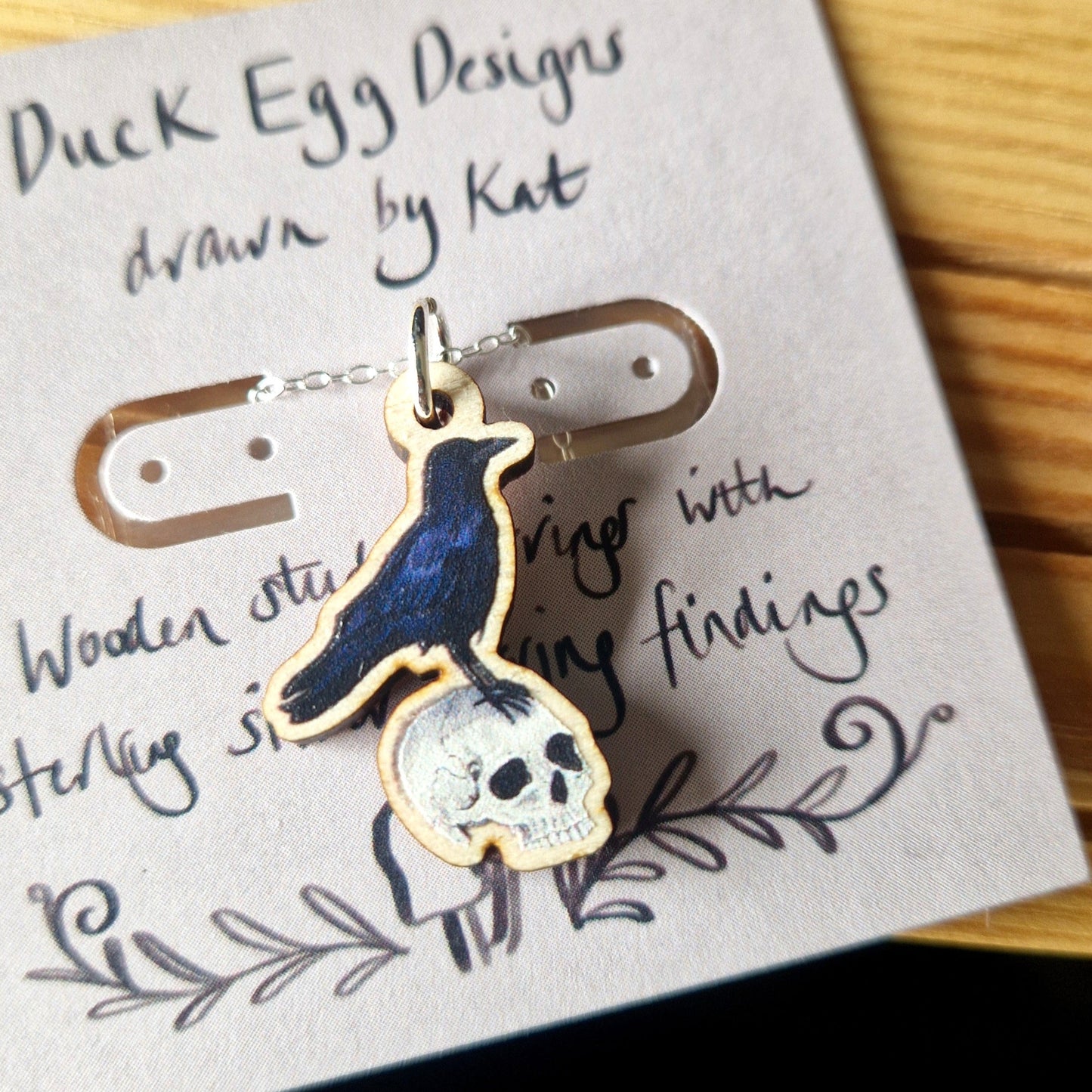 Crow on a human skull jewellery pendant on a silver chain on a card with 'Duck Egg Designs' text.
