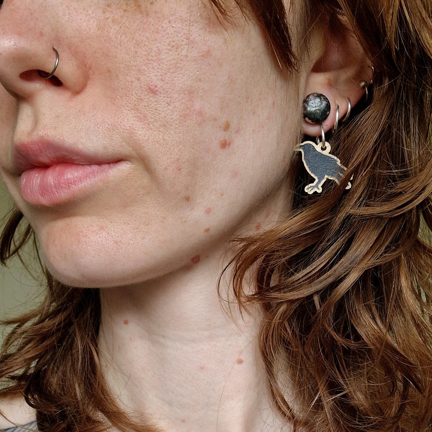Close-up of a person wearing raven earrings and a silver nose ring.