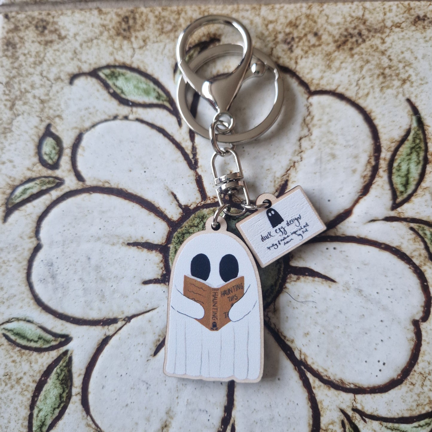 The Reading Ghost Keyring from Duck Egg Designs features a cute cartoon ghost reading "Haunting 101" and a mini notepad charm, shown on a floral-patterned surface.