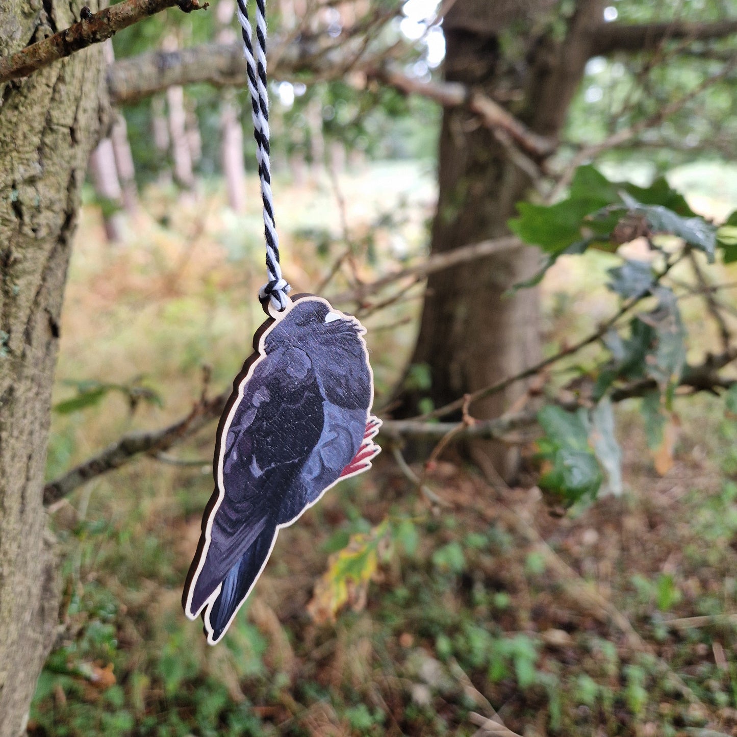 The Bob Chonky Pigeon Hanging Decoration by Duck Egg Designs hangs from a black-and-white string on a tree branch in a wooded area with green foliage in the background.